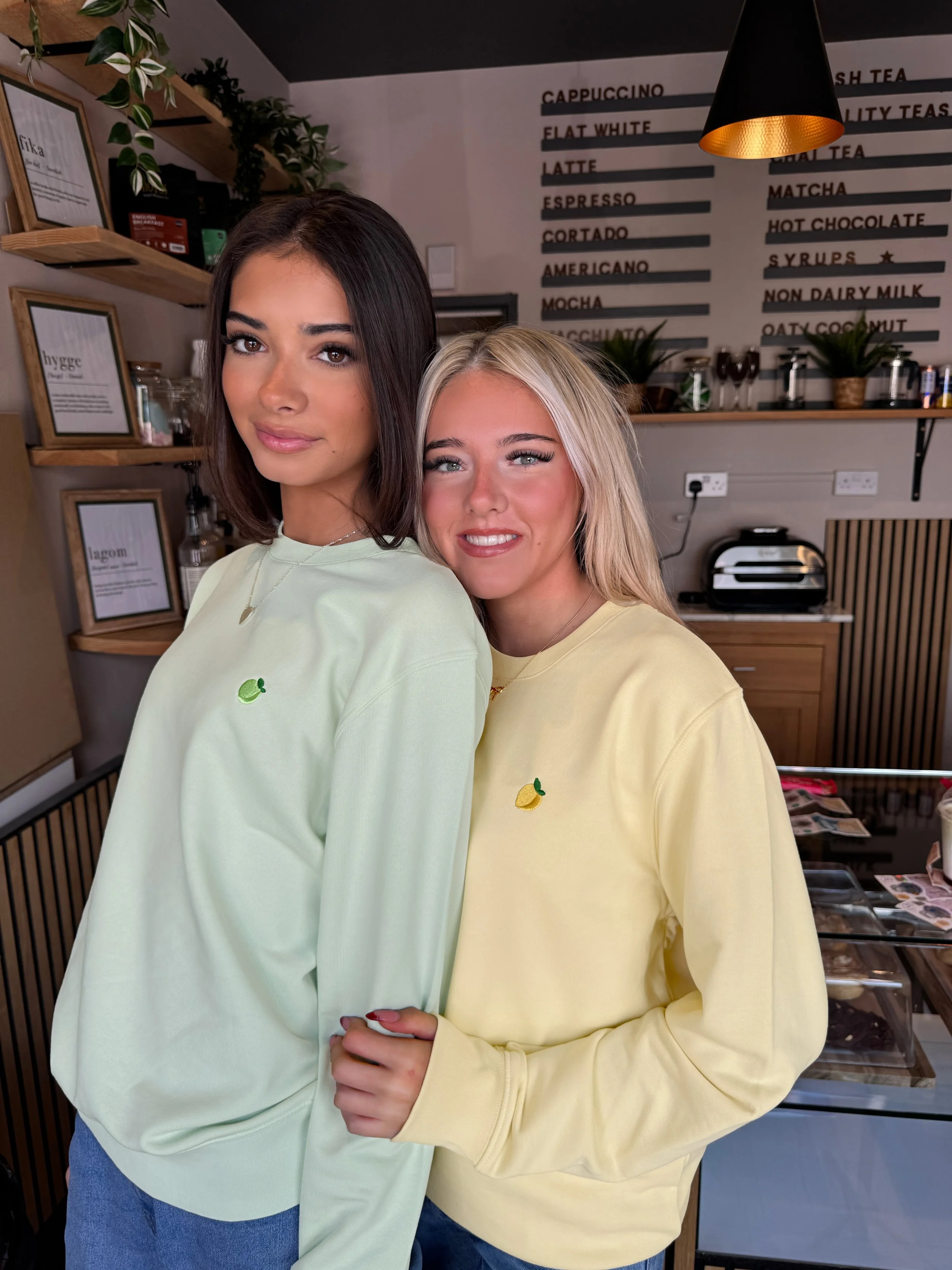 Two women standing close together inside a cafe, wearing pastel yellow and green sweatshirts with small embroidered lemon designs, smiling at the camera.
