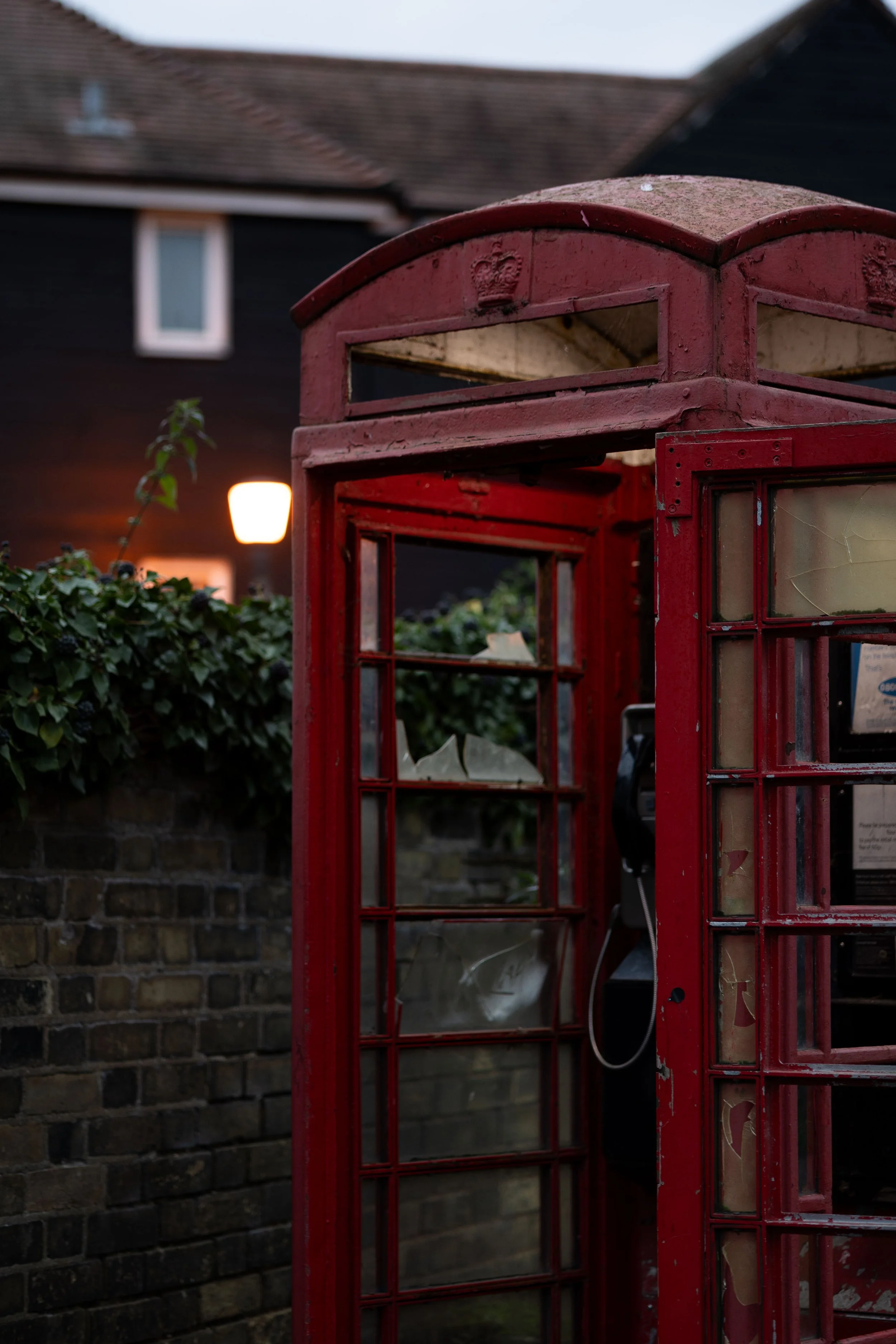 An old, weathered red British telephone booth with broken glass, situated outdoors near a brick wall and greenery.
