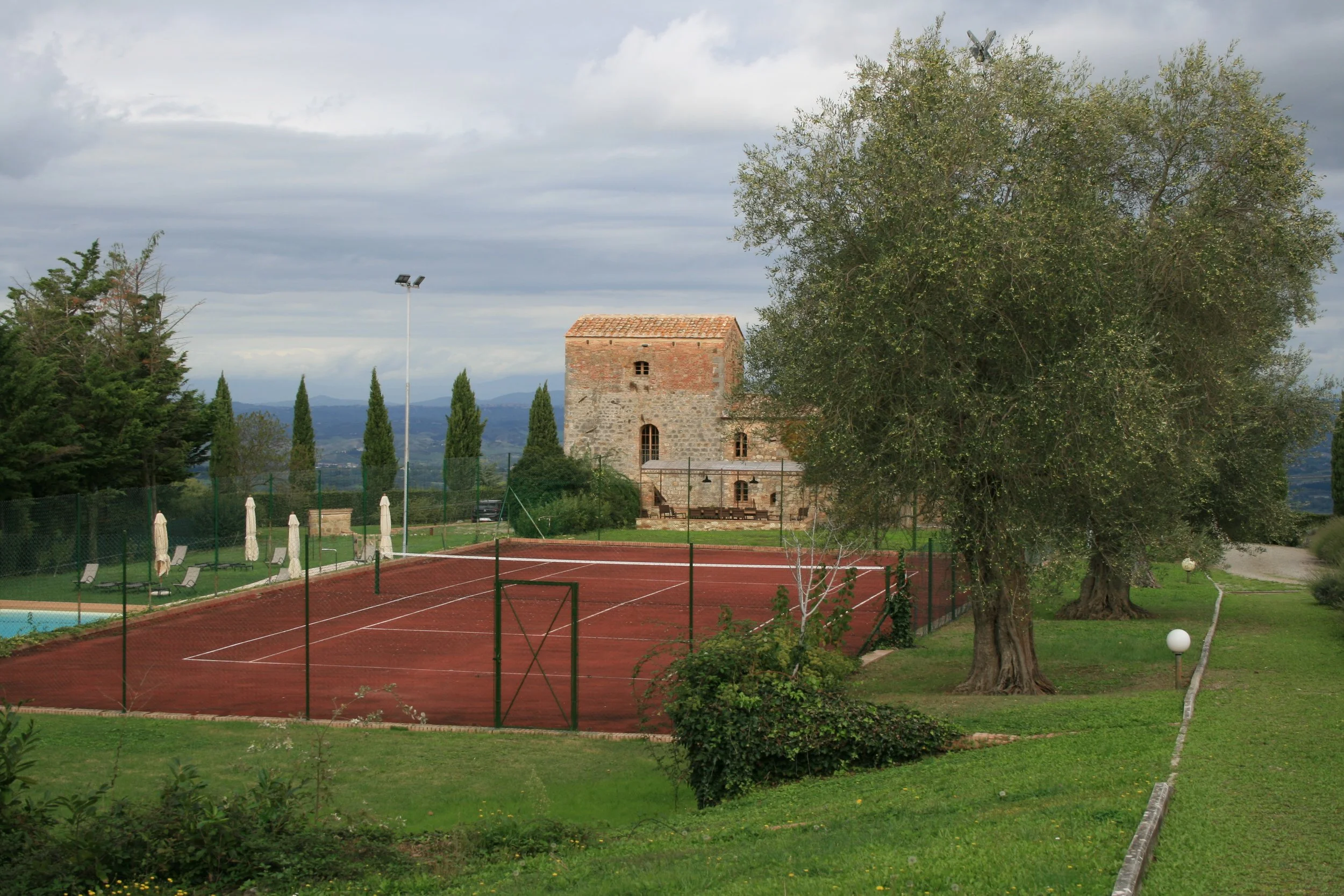 A tennis court with red clay surface, surrounded by a black fence and white parasols, with a historic stone tower in the background, large trees on the right, and cloudy sky overhead.