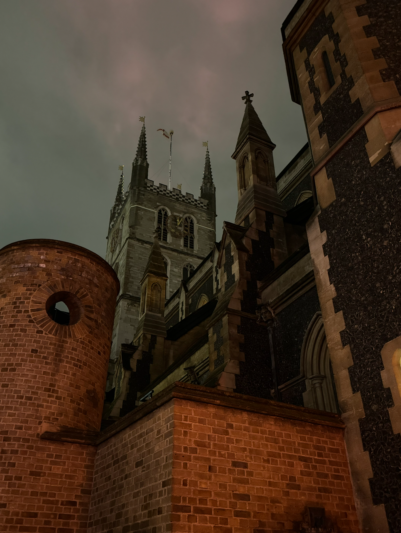 Nighttime view of a historic brick church with a tall clock tower and pointed spires, illuminated subtly against a cloudy sky.