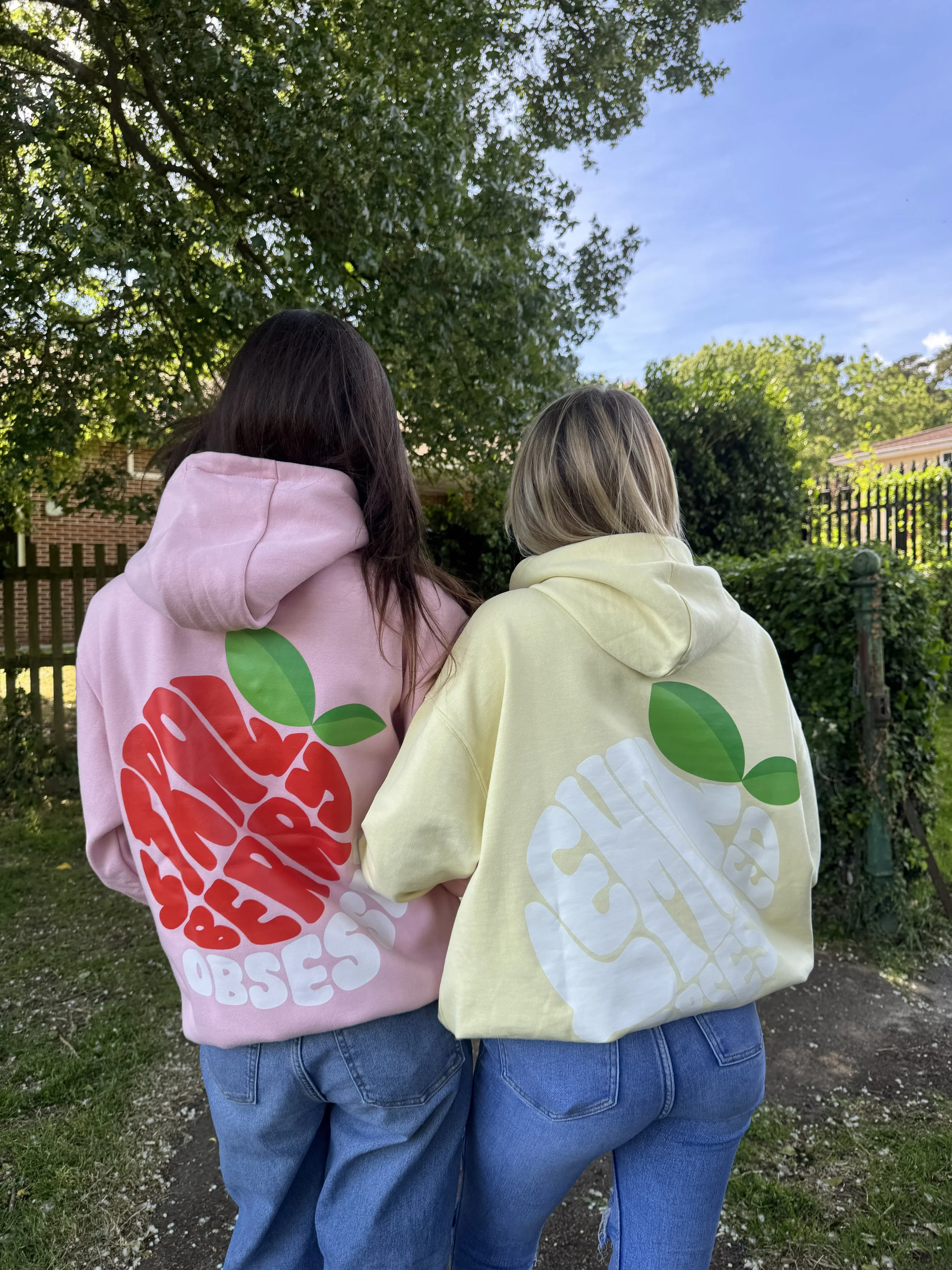 Two women, seen from behind, are standing outdoors under a tree. They are wearing hoodies with large fruit-themed graphic designs on the back, one pink with a red and green design, and the other yellow with a white and green design. Both are wearing 