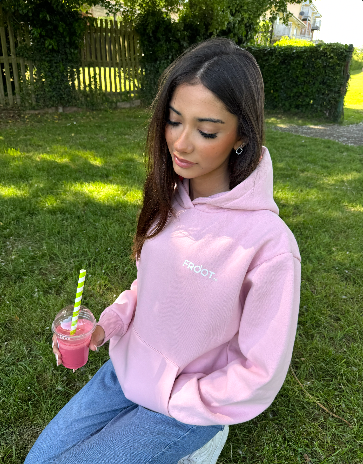 Young woman with long brown hair wearing a pink hoodie with 'FROOT Co' logo, sitting on grass in a park, holding a pink smoothie with a green-striped straw, surrounded by greenery and a wooden fence.