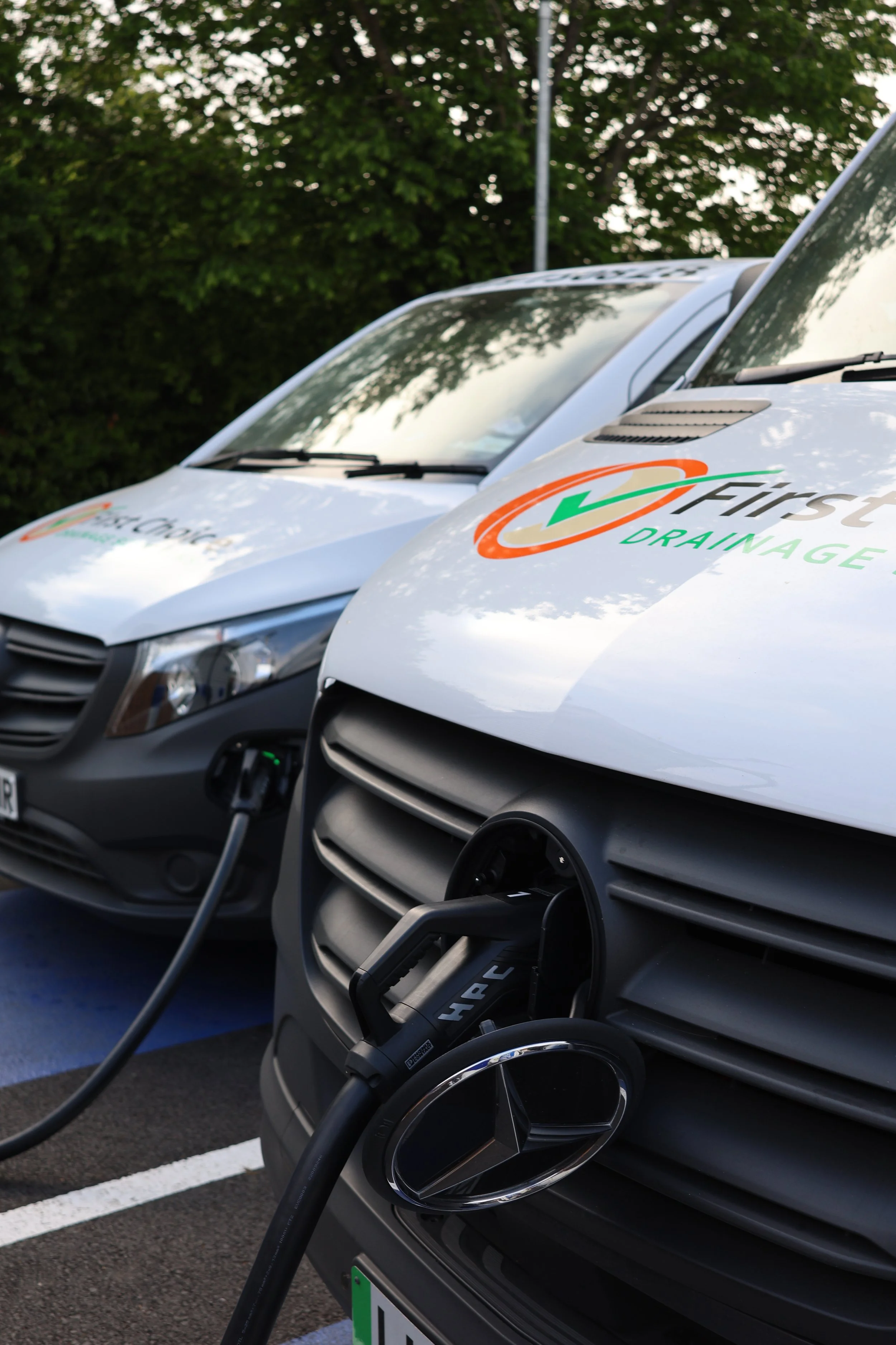 Two white electric delivery vans with logos reading 'First Drainage' are parked outdoors, connected to charging stations.