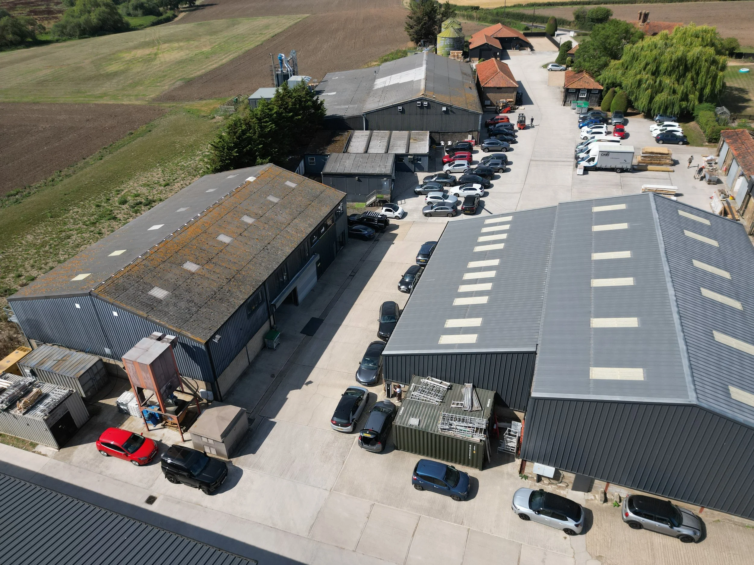 Aerial view of an industrial area with large metal buildings, parked cars, shipping containers, and surrounding farmland.