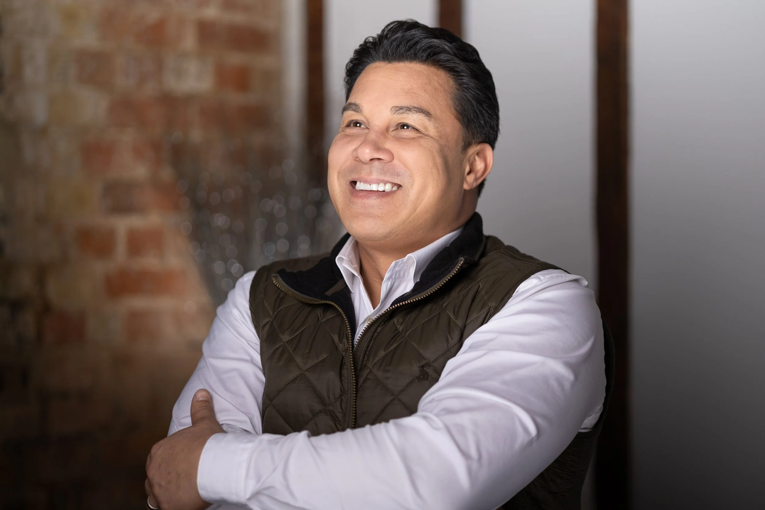 A smiling man with dark hair, wearing a white shirt and a black quilted vest, standing with arms crossed in front of a brick wall and a blurred background.