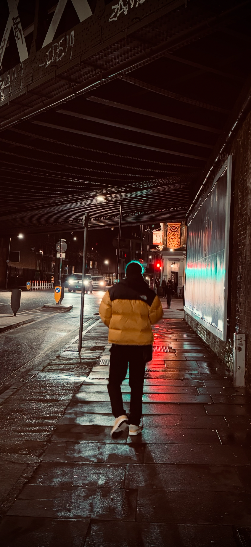 A person walking on a wet city sidewalk at night, under a bridge, with cars passing by and traffic lights in the background.