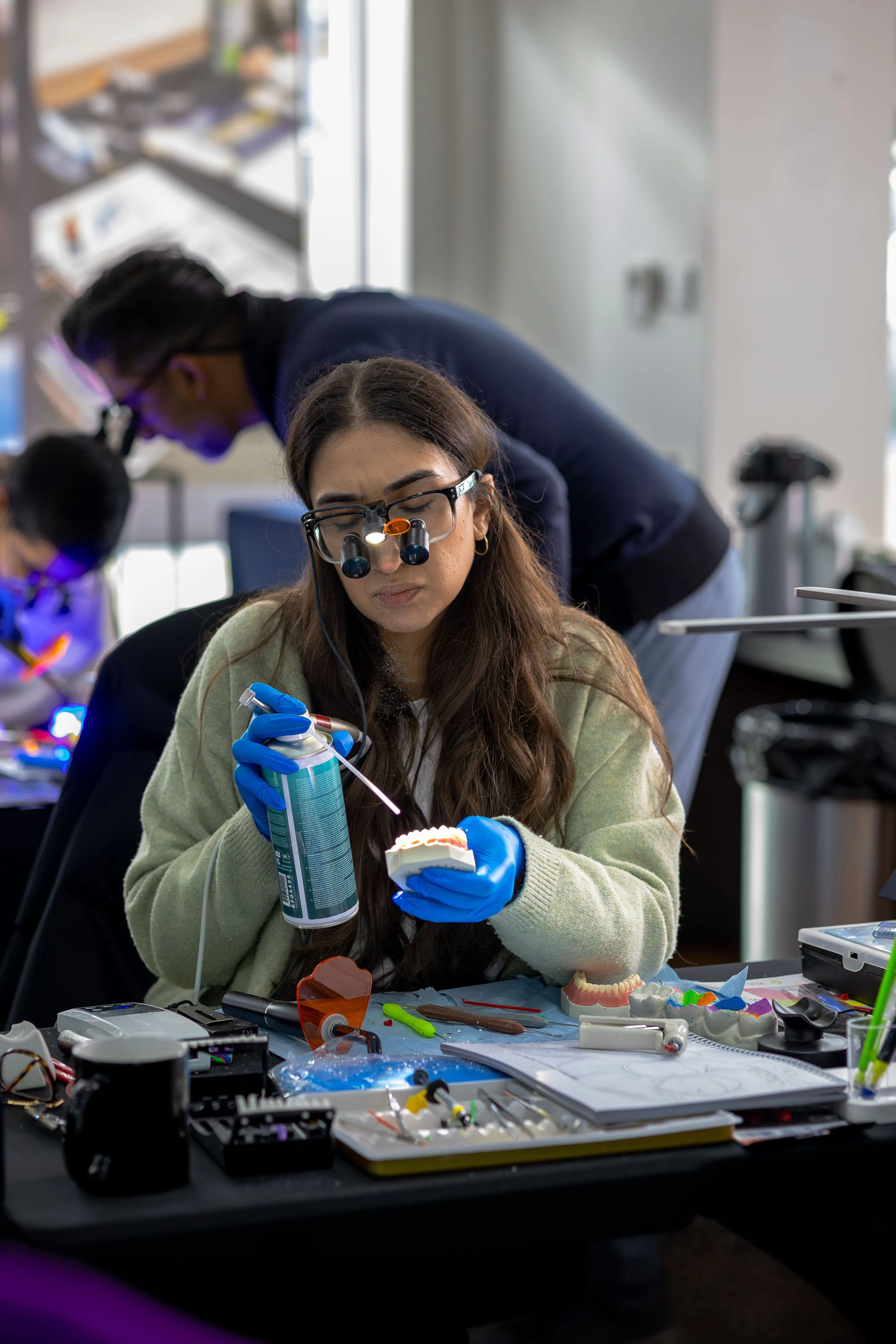 A woman wearing magnifying glasses with lights is working on a dental model with a syringe, surrounded by dental tools and materials on a cluttered table, in a laboratory or classroom setting with other people working in the background.