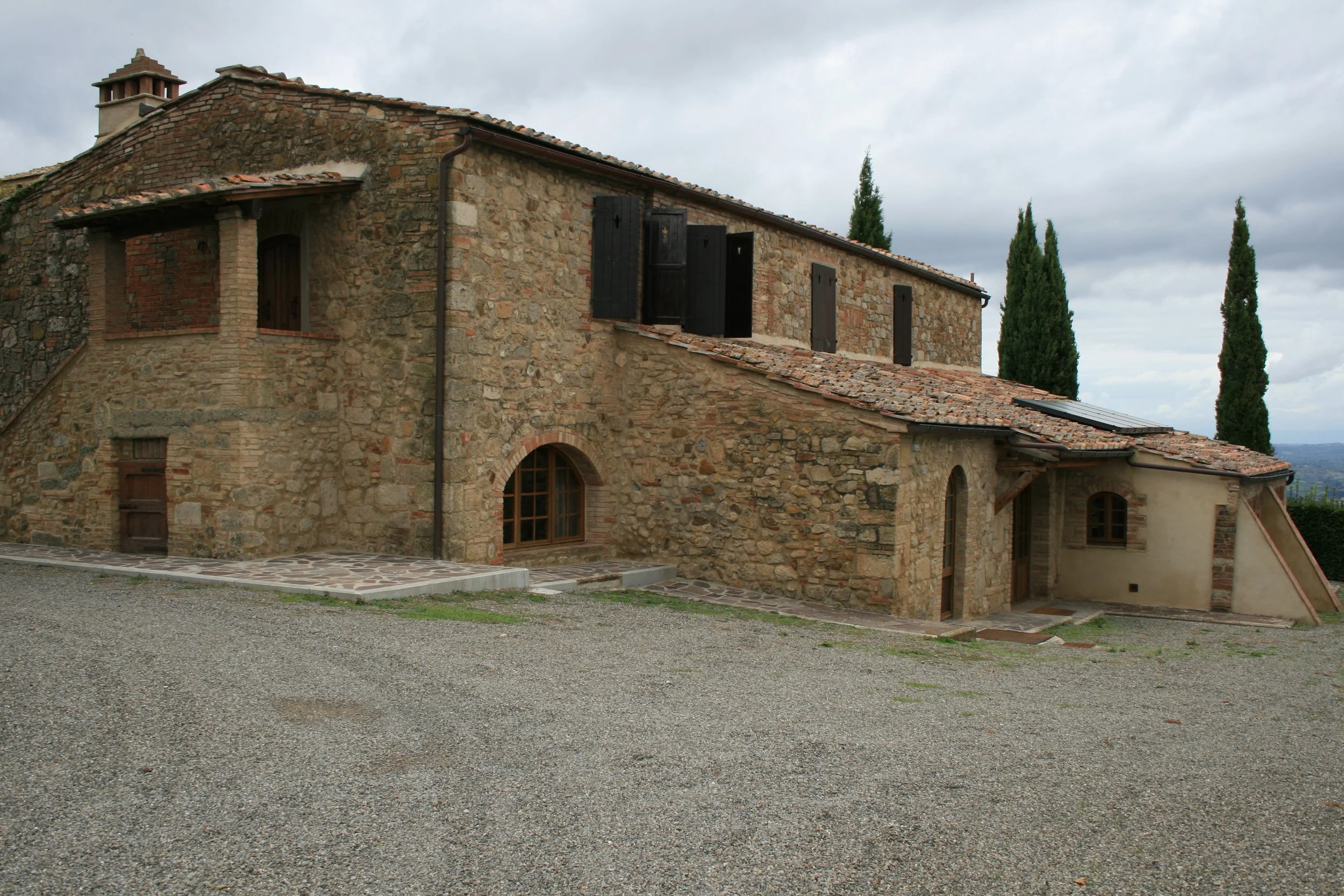 A rustic stone house with multiple levels, arched doorway, and black window shutters, surrounded by tall cypress trees under a cloudy sky.