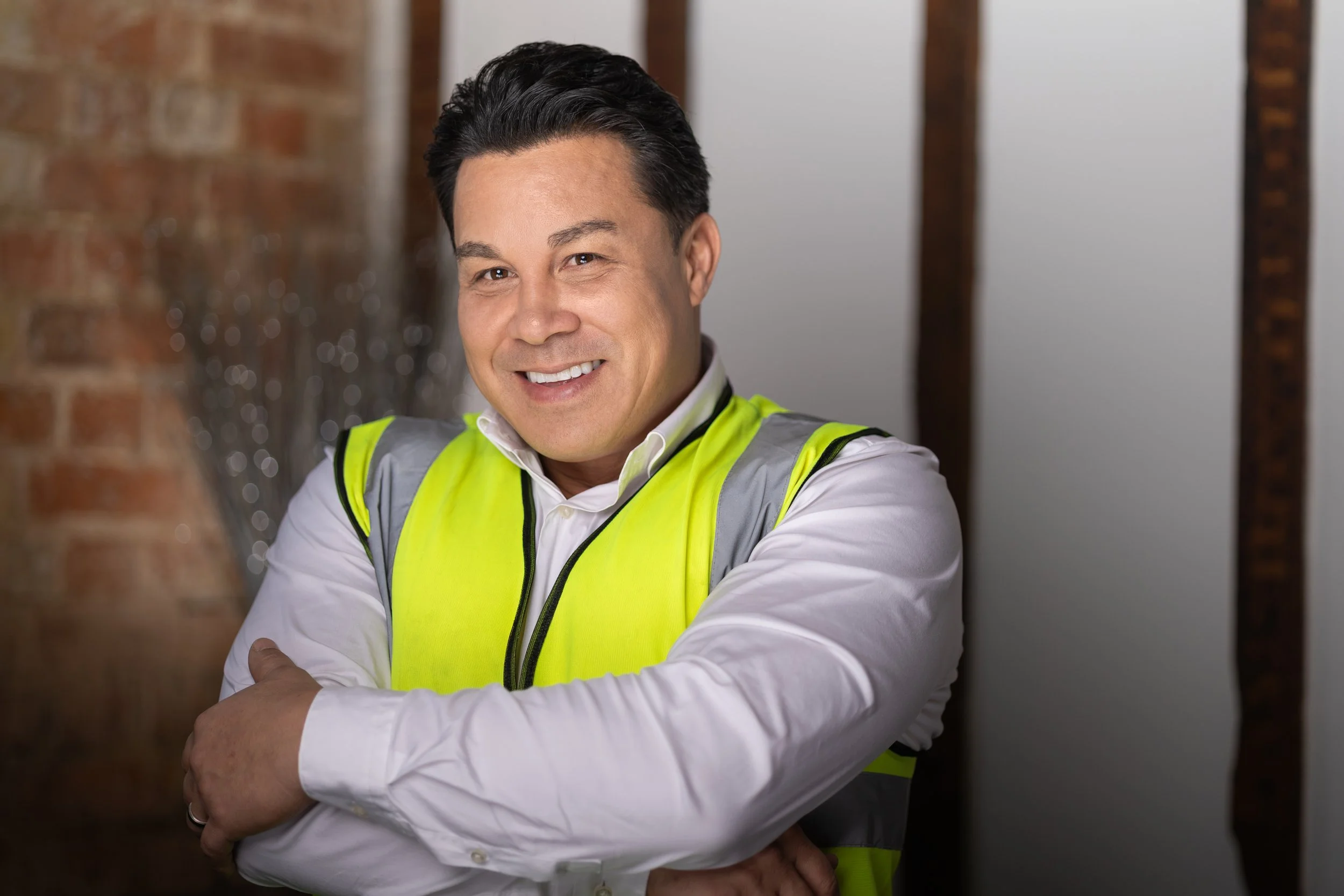 A smiling man in a white shirt and yellow safety vest standing indoors with arms crossed, brick wall on the left, and wooden beams on the right.