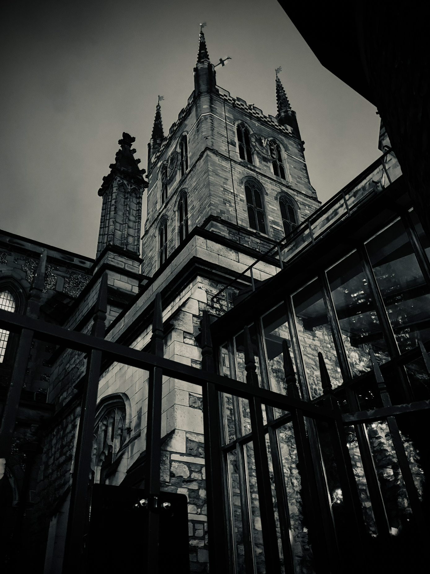 A black and white photograph of a historic church tower with gothic architecture, stone construction, and spires reaching upward, taken from a low angle with metal railing in the foreground.