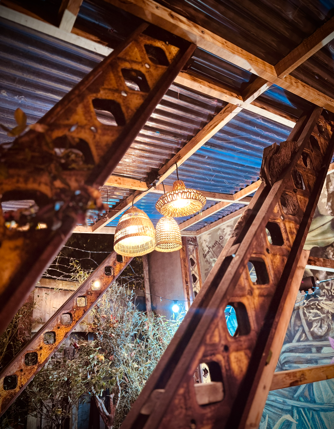 Interior view of a space with a corrugated metal ceiling, wooden beams, hanging woven pendant lights, rustic metal stairs with cutout patterns, and plants outside visible through a window.