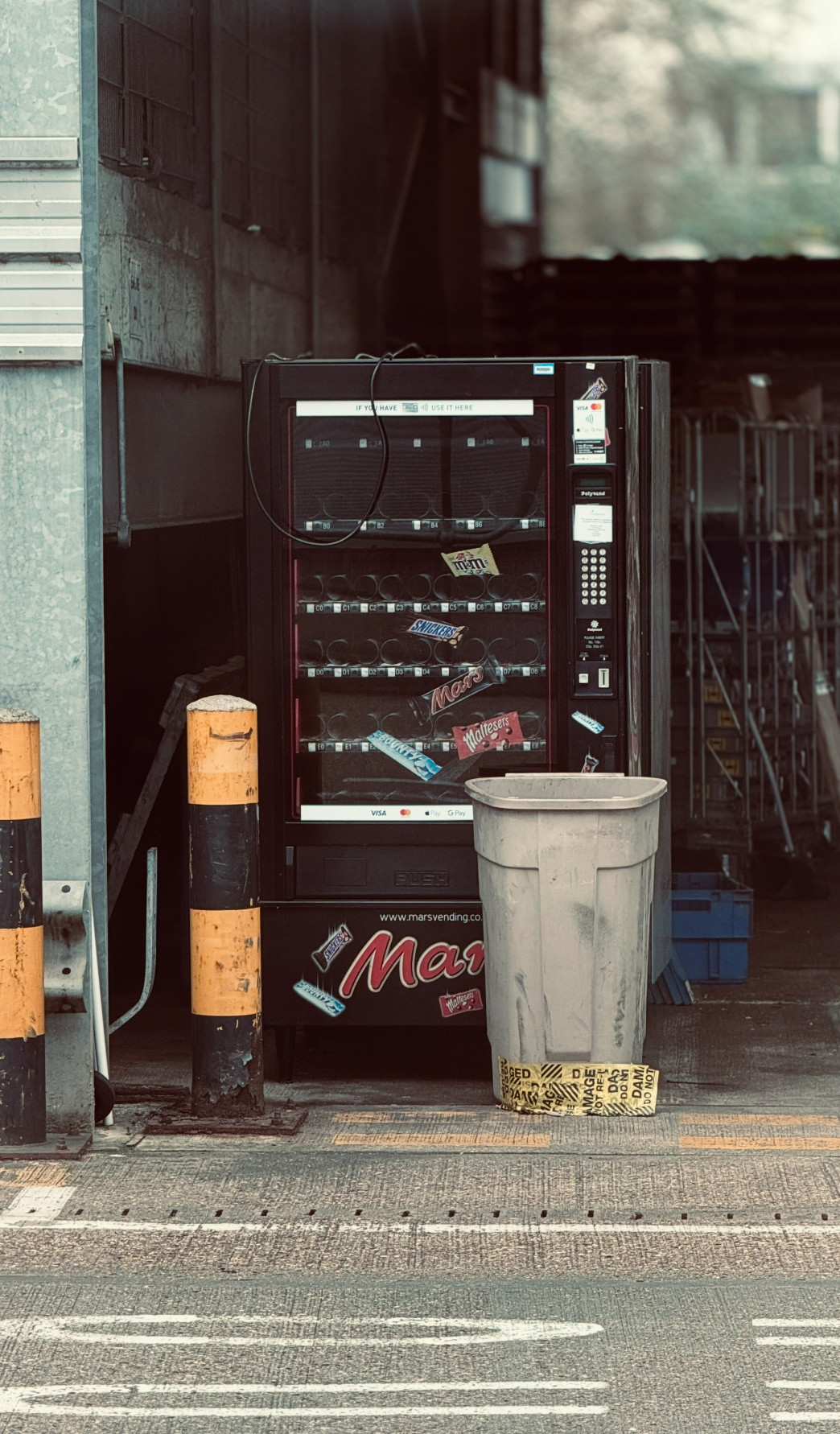 A vending machine outside a store with candy packaging on and below it, and a large trash bin in front with caution tape around it. Two orange and black safety bollards are on the left side of the vending machine.