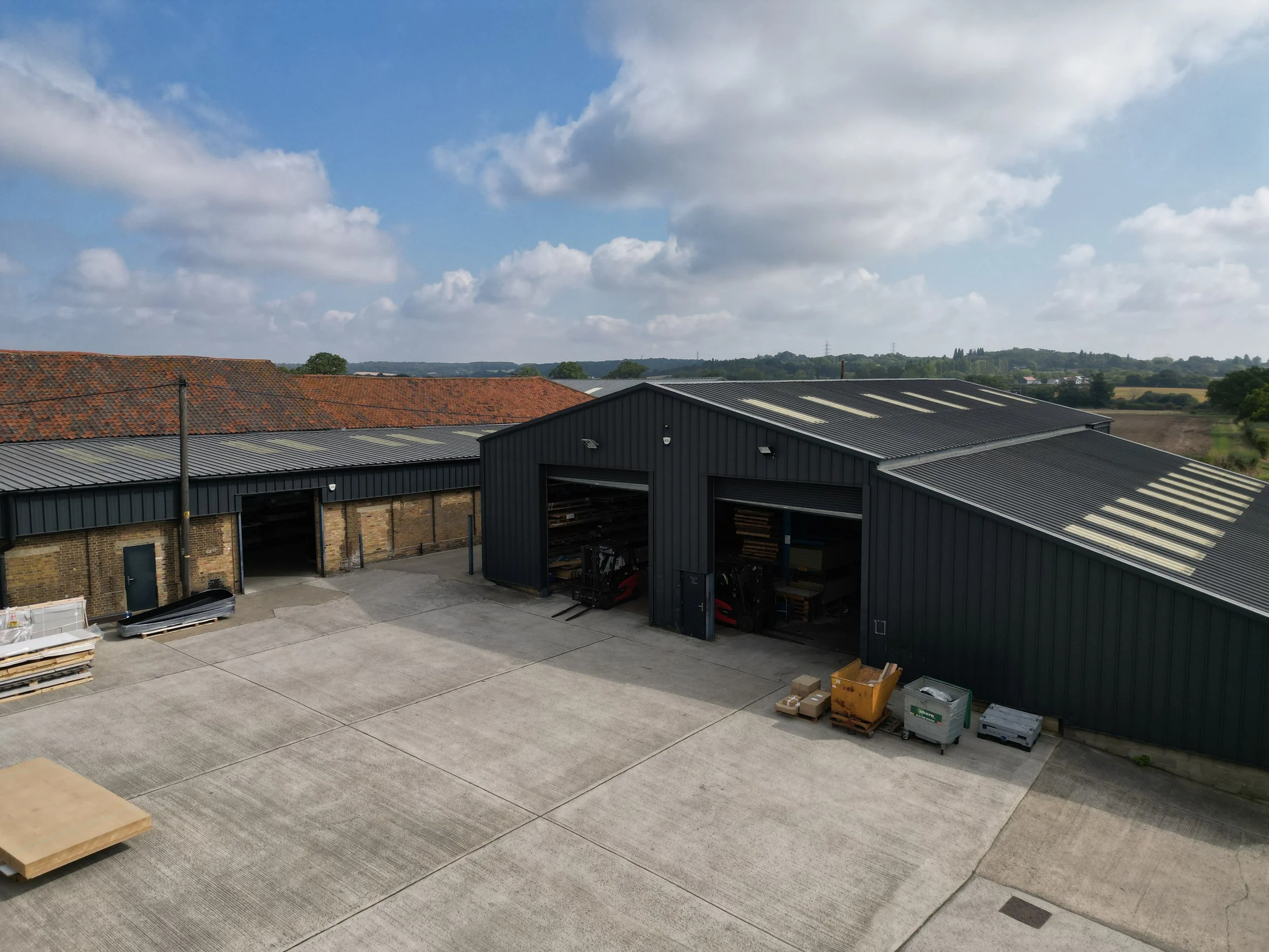 Large industrial warehouse with black metal siding, open doors revealing forklifts, items, and shelves inside, alongside a paved outdoor area with pallets, carts, and storage containers. Background shows partly cloudy sky and distant countryside.