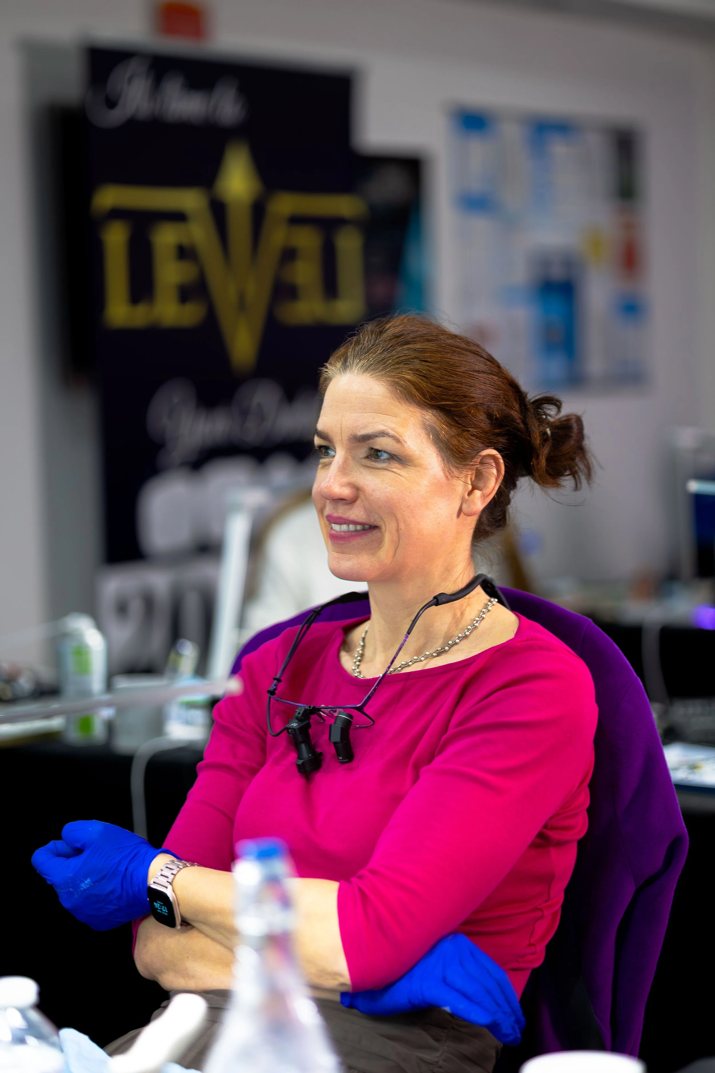A woman with red hair wearing a pink shirt, blue gloves, and a watch, smiling while seated at a table with medical or laboratory supplies, in a room with posters and banners on the wall, including one that says 'Hard to LEV'.