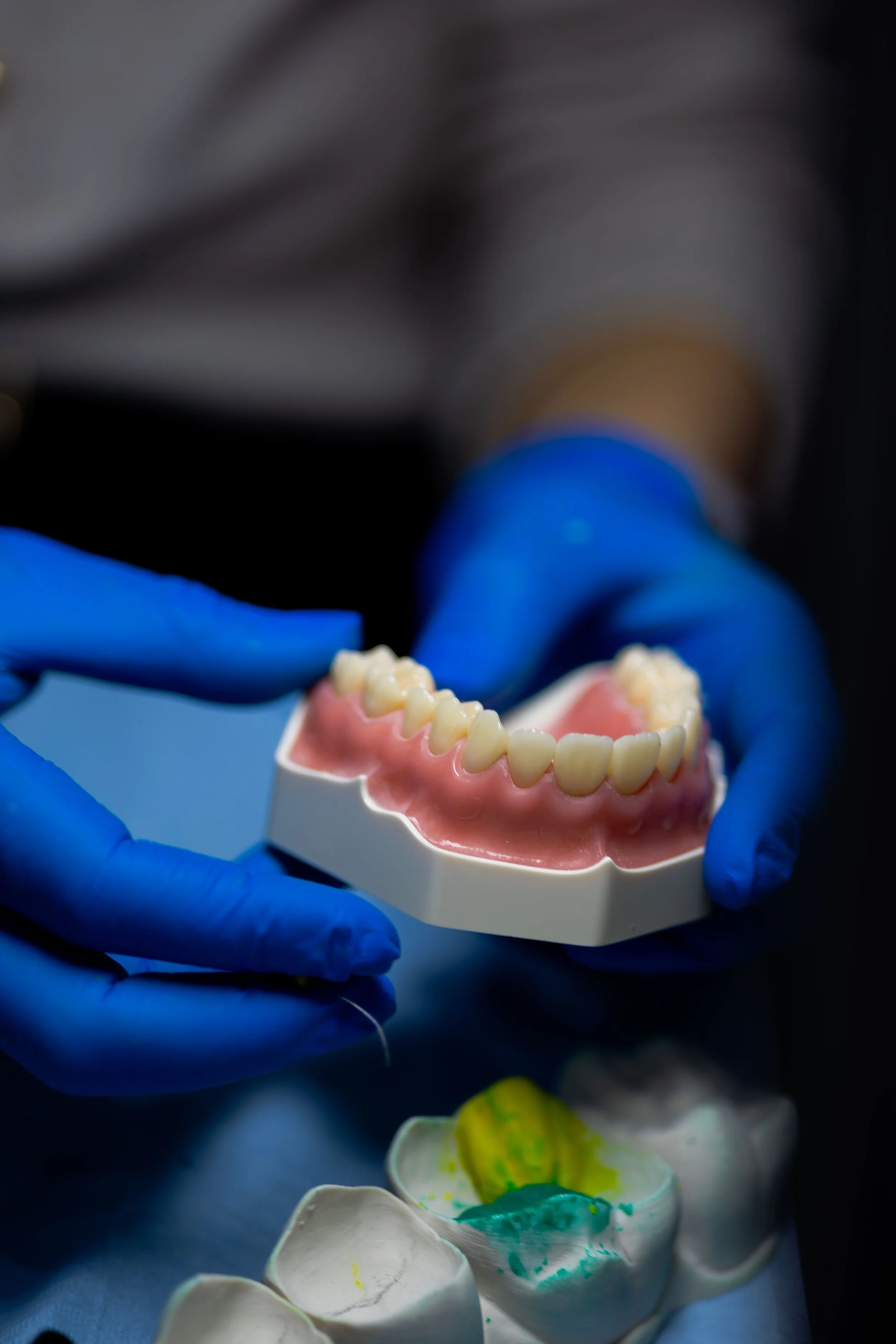 Close-up of a dental model of teeth held by a person wearing blue gloves, with dental tools and colorful dental materials in the background.