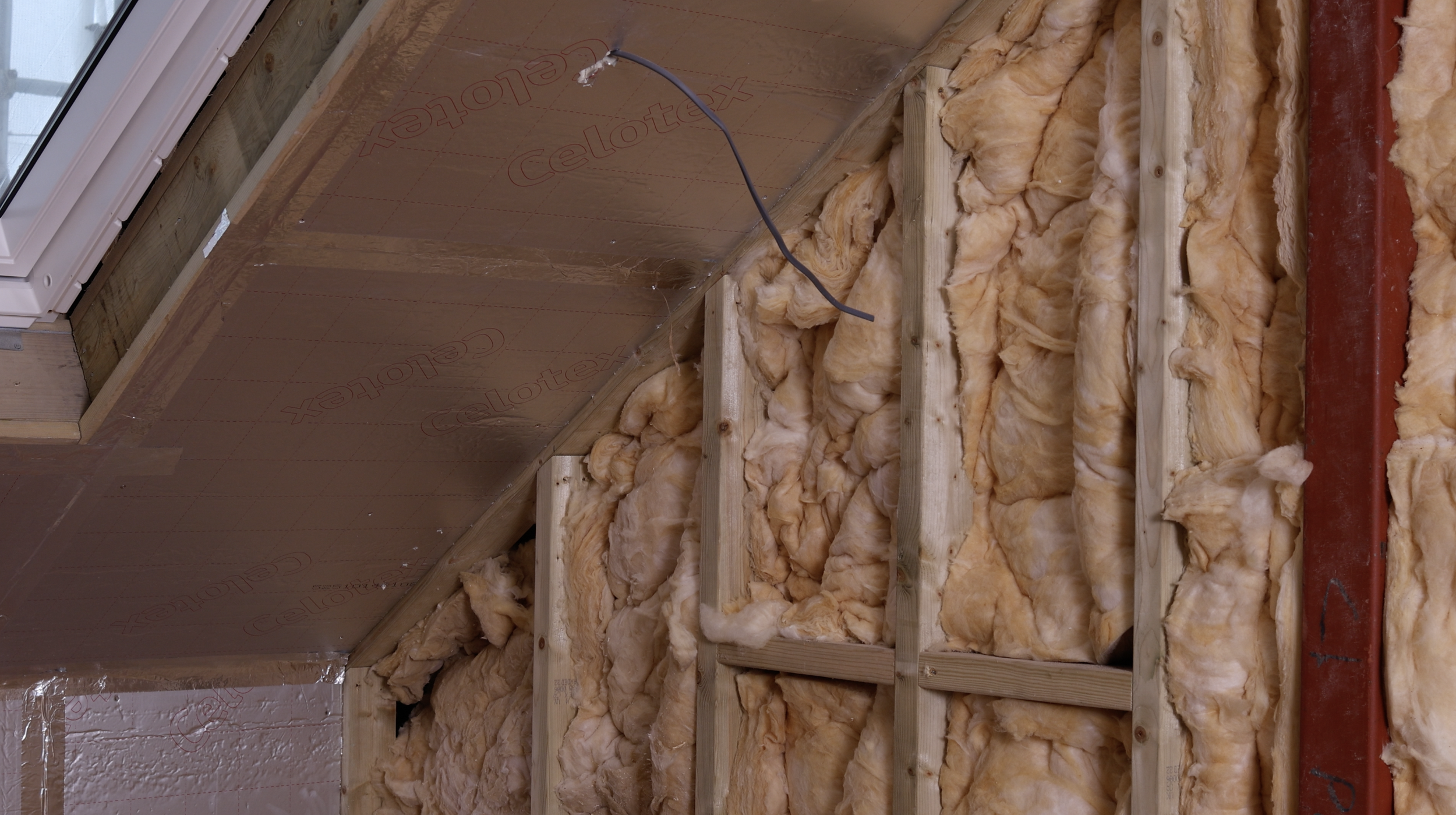Interior of an unfinished attic or wall with insulation, wooden framing, a power cord hanging, and a red metal support beam.