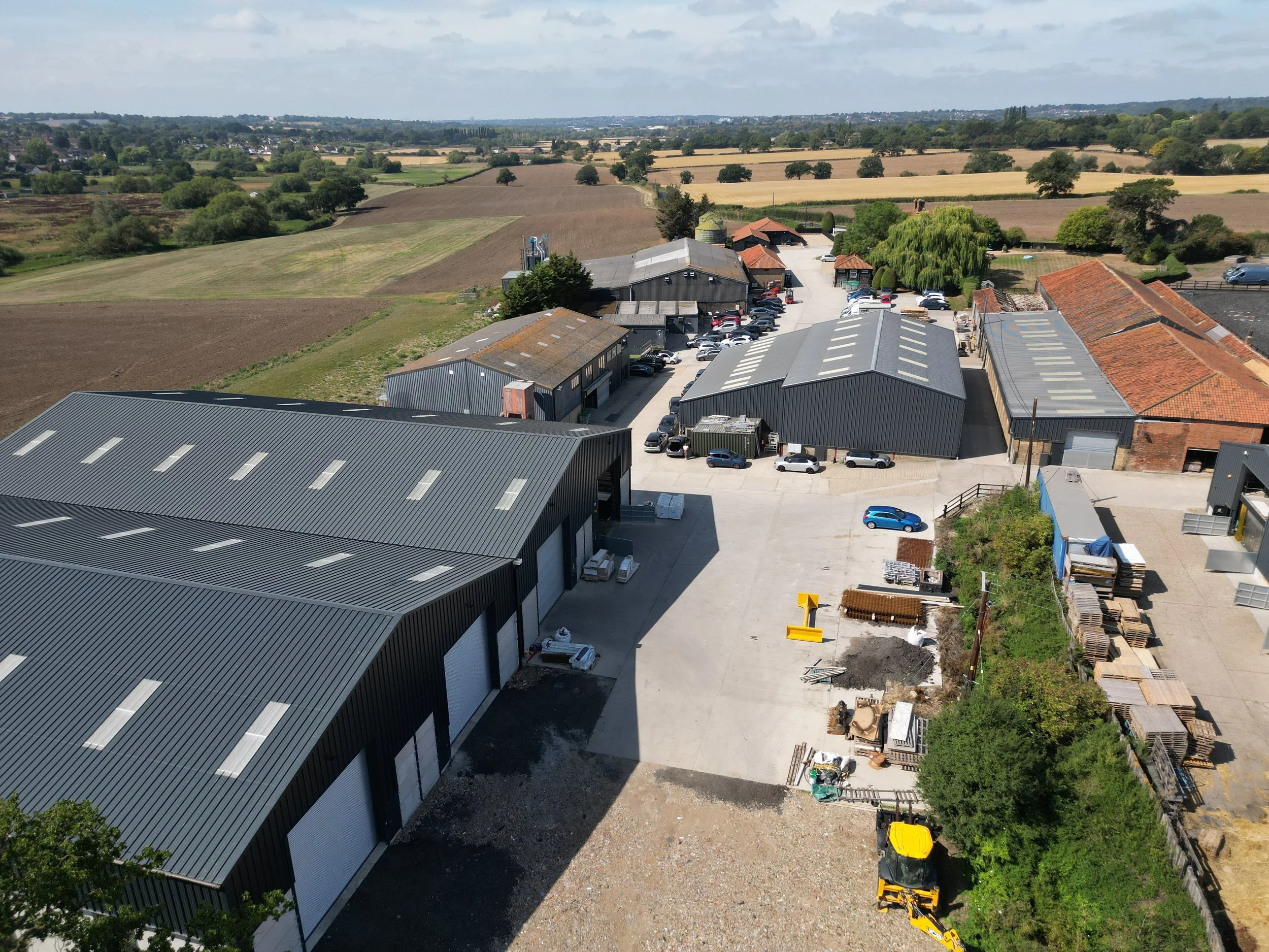 An aerial view of an industrial area with multiple metal warehouses, parked cars, and construction materials. Surrounding farmland is visible in the background.