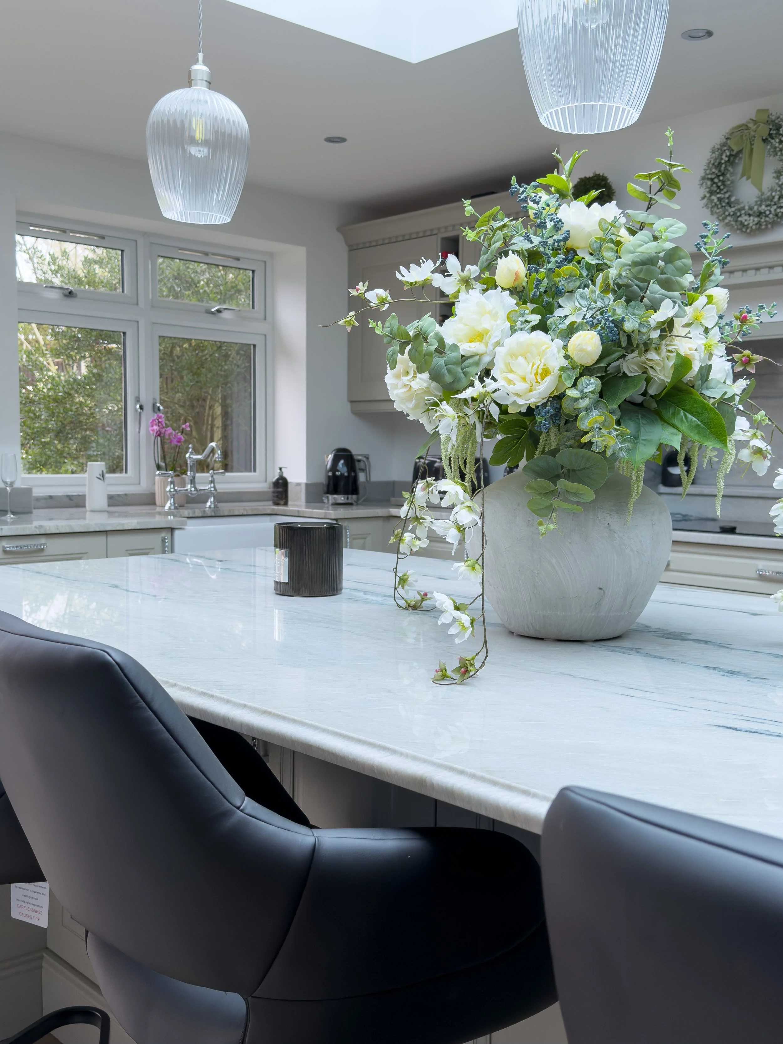 A modern kitchen with a white marble island and black chairs, decorated with a large floral arrangement in a white ceramic vase, featuring white flowers and green foliage. Two pendant lights hang from the ceiling, and a window shows outdoor greenery.