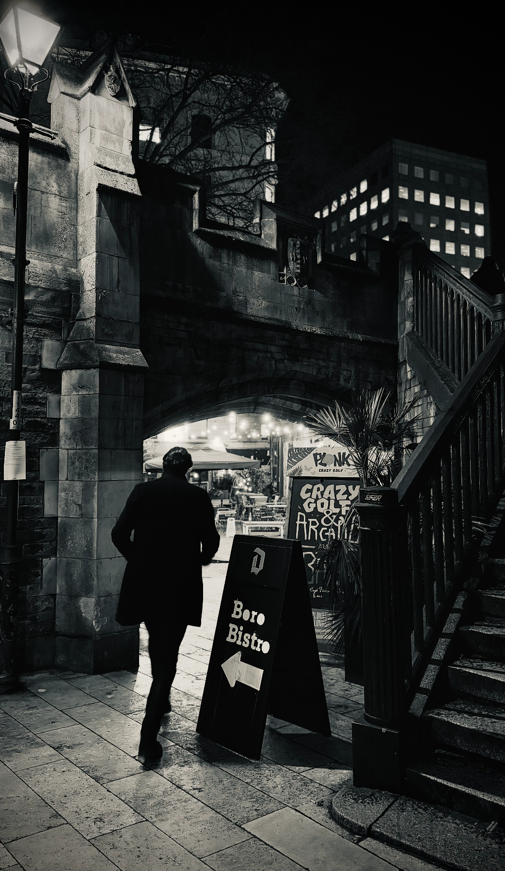 A man dressed in dark clothing walking past a blackboard sign that reads 'Boro Bistro' with an arrow pointing left. The scene is at night outside an urban bistro or restaurant, with an arched stone bridge overhead, street lamps, some outdoor seating,