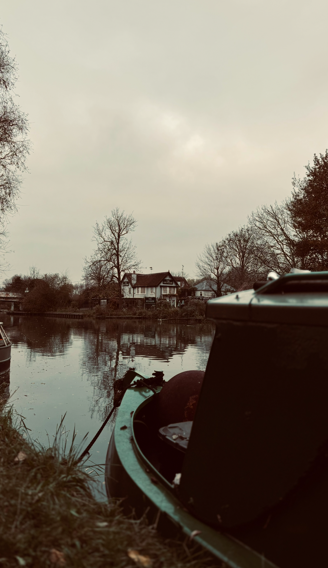 View of a calm river with a house and trees on the opposite bank, partially obscured by a boat moored at the riverbank in the foreground, under an overcast sky.