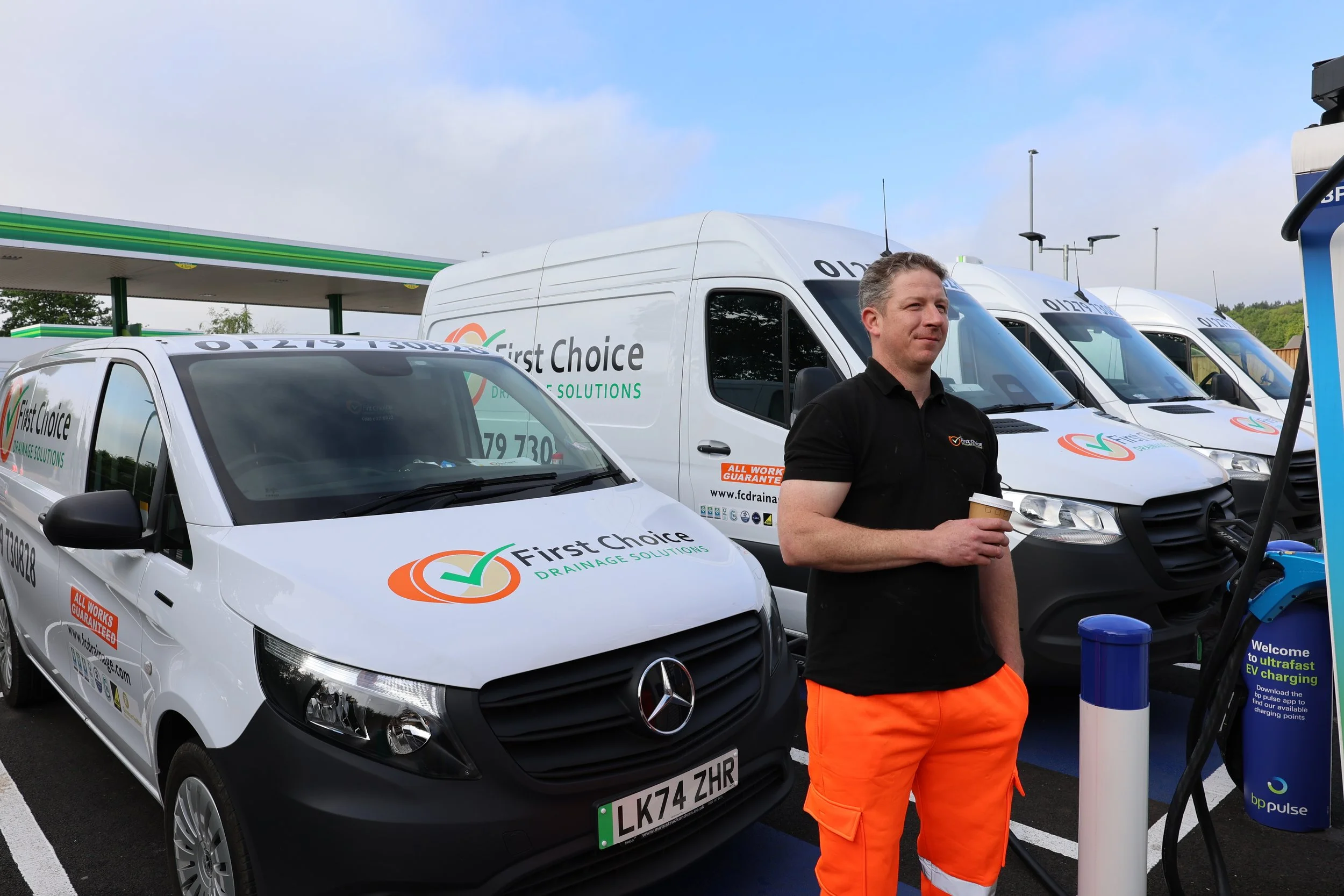 A man standing at an electric vehicle charging station with three First Choice Drainage Solutions work vans parked behind him. The man is holding a coffee cup and wearing black shirt and orange pants. The vans display the company's logo and contact i