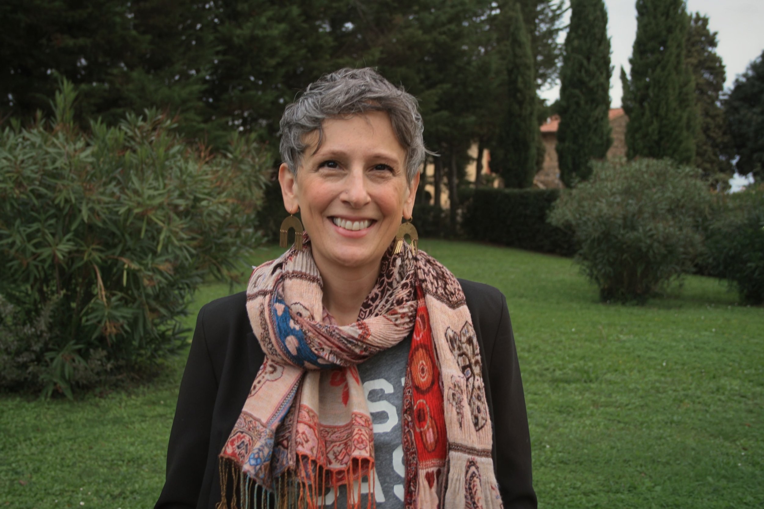 A smiling woman with short gray hair and large earrings, wearing a patterned scarf and black jacket, standing outdoors in a garden with green grass, trees, and bushes.