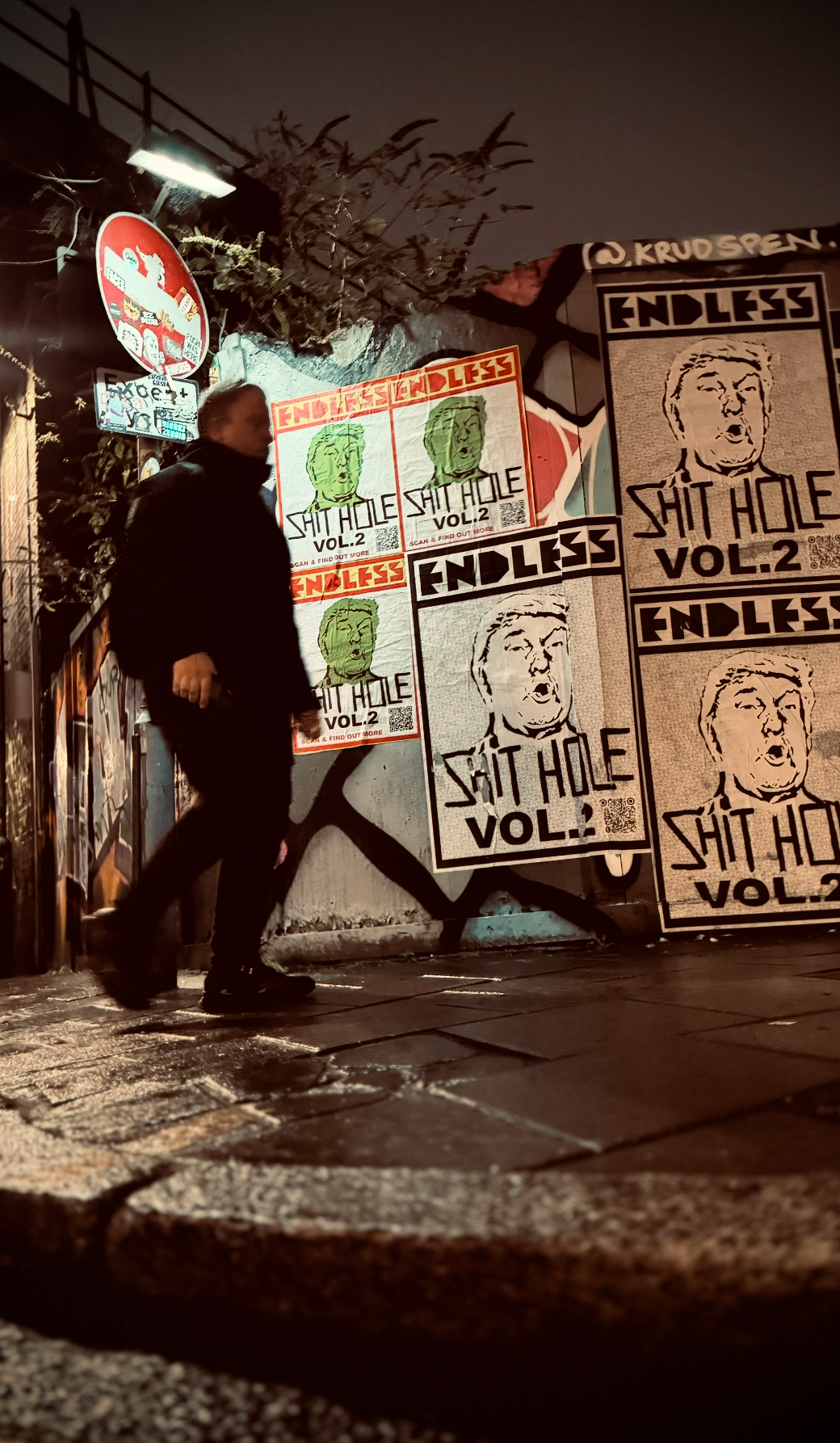 Street scene at night with posters on a wall. Posters display the face of Donald Trump with the text "Endless Sit Hole Vol.2" and a colorful illustration of Donald Trump with the same text. A person dressed in dark clothing walks past the posters, pa