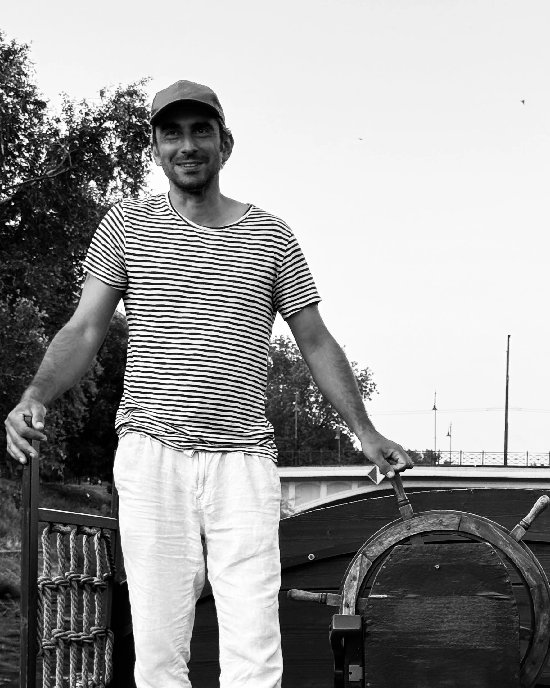 A man in a striped t-shirt and light-colored pants standing outdoors next to a wooden ship's wheel, holding a stick or handle, with trees and a bridge in the background.