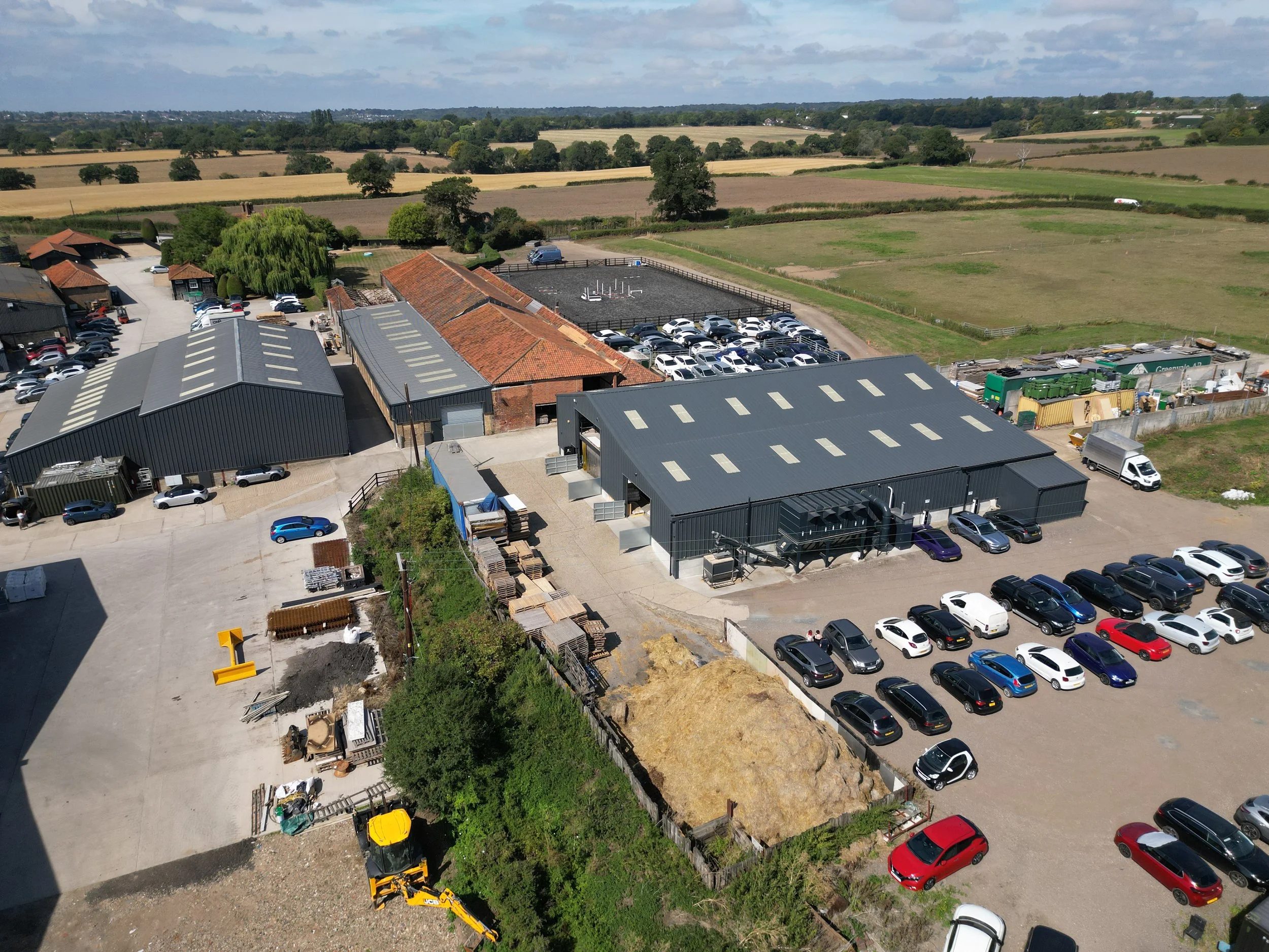 Aerial view of a farm or rural property with multiple buildings, parking lots filled with cars, surrounding fields, and a construction site with machinery.