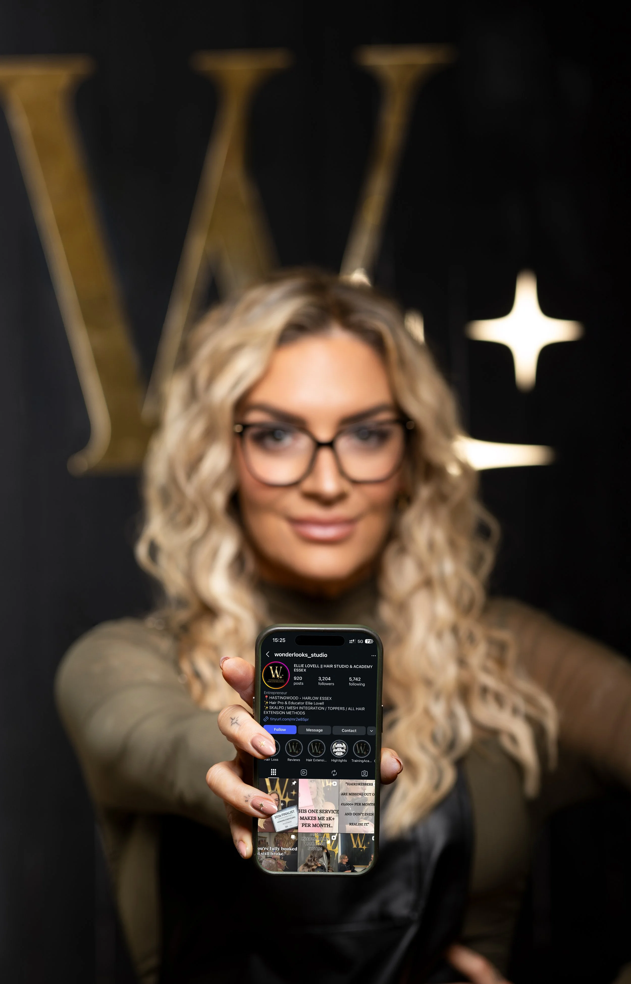 A woman with blonde curly hair and glasses holding a smartphone showing an Instagram profile, standing in front of a black background with a large gold 'W' logo.