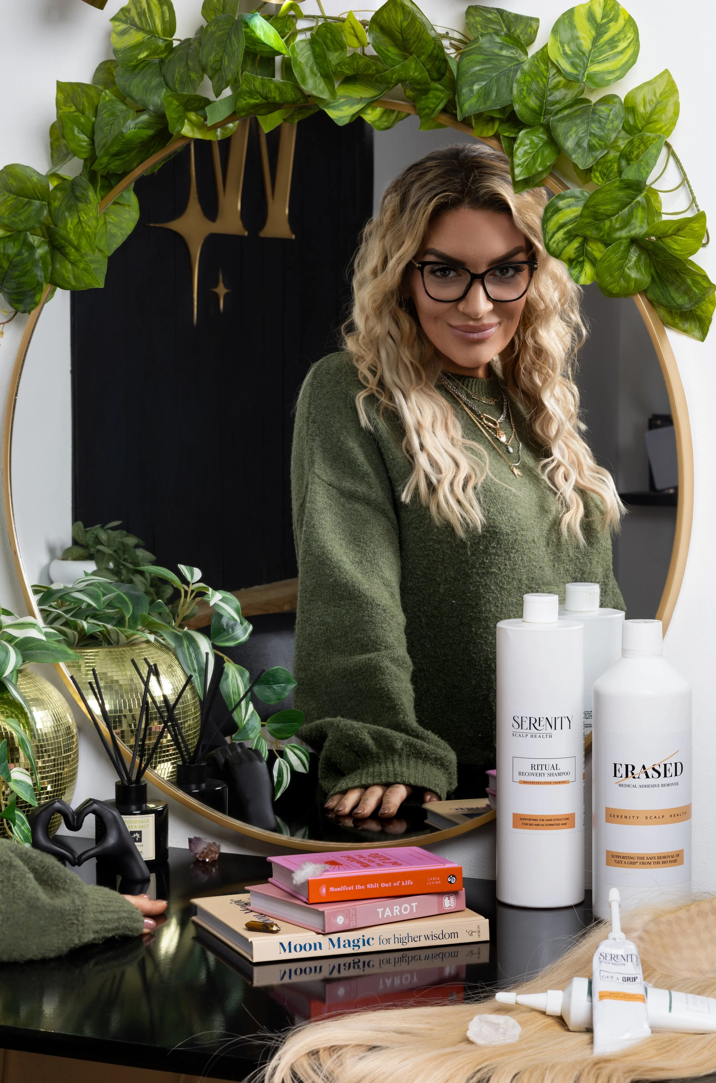 A woman with long curly blonde hair, glasses, and a green sweater looking into a mirror surrounded by plants and skincare products, including shampoo, an adhesive remover, and tarot cards on a black table.