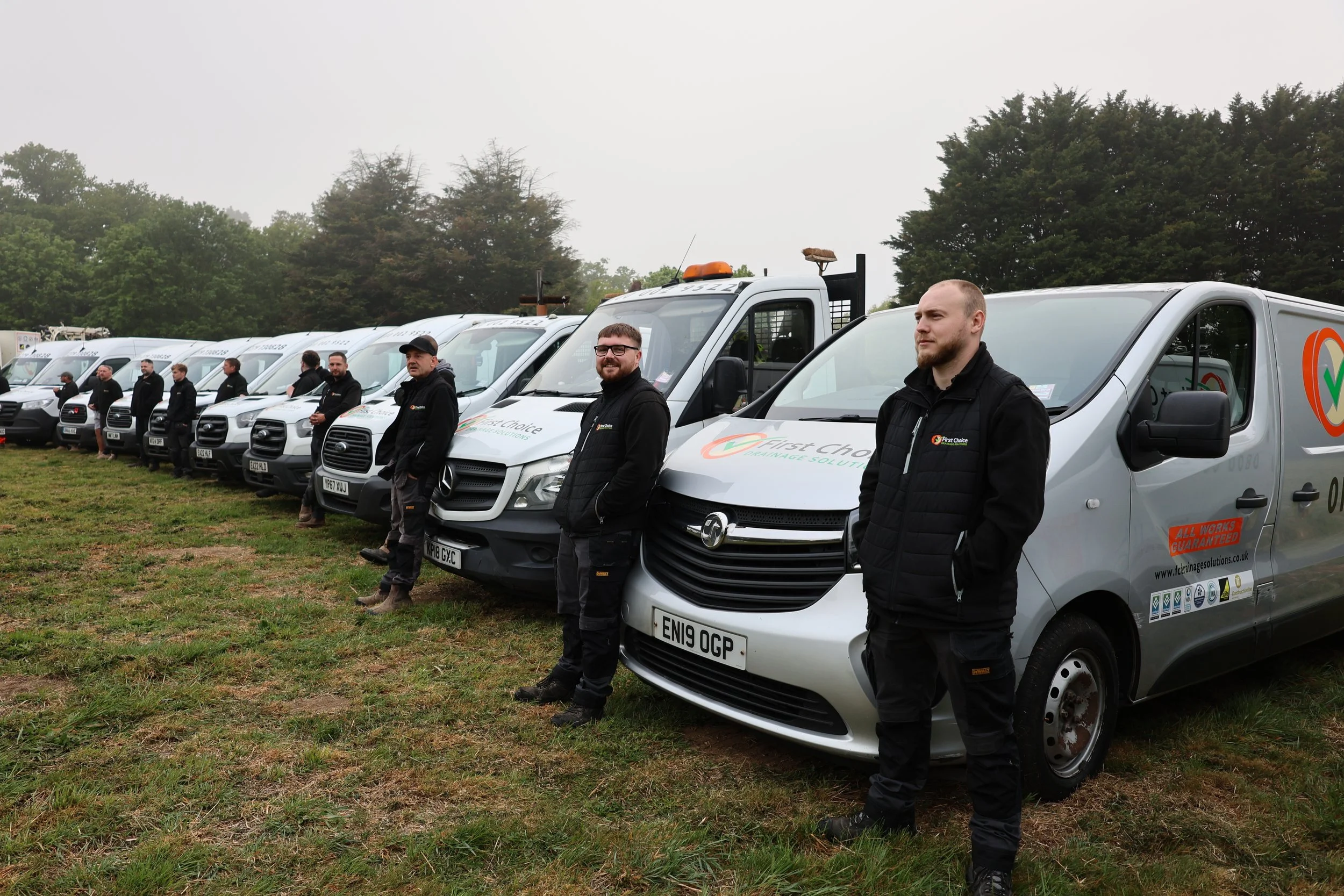 Line of white company vans parked on grass with employees standing next to each vehicle, wearing black jackets, some smiling and some looking serious, with trees and fog in the background.