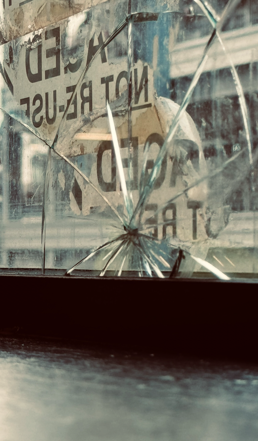 Close-up of broken glass in a shattered window with a 'Do Not Block' traffic sign reflected in it.