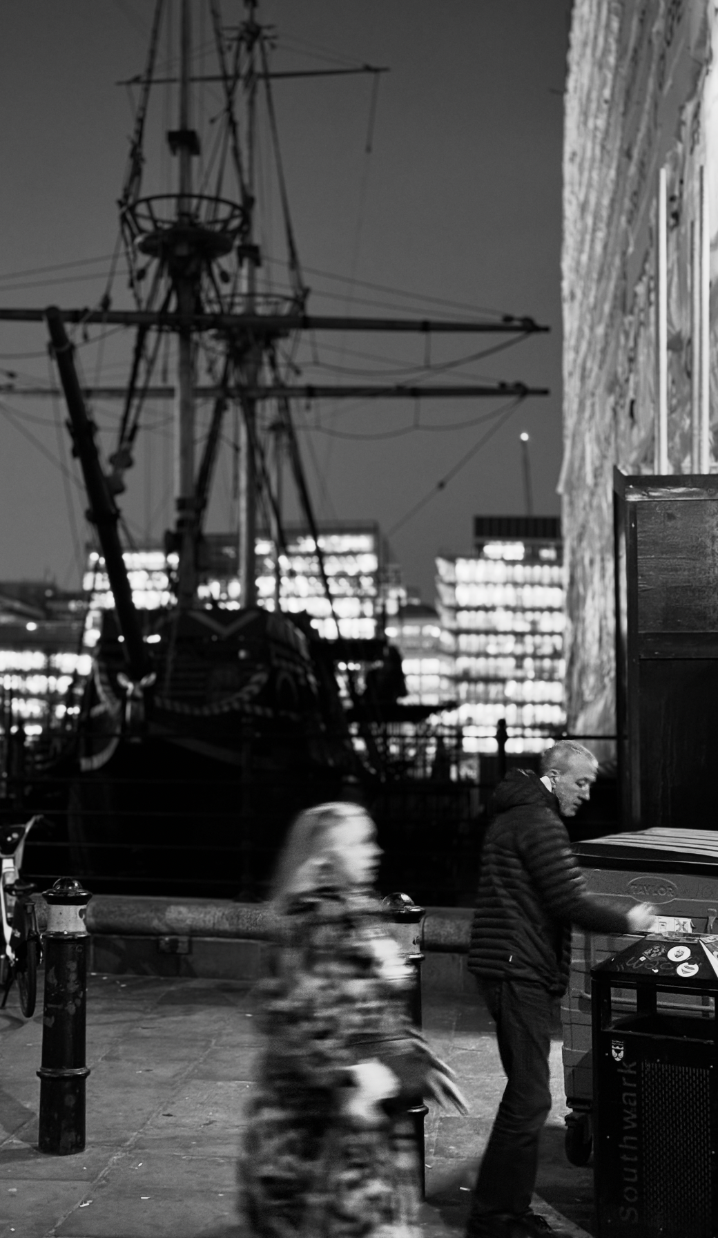 Night scene at a harbor with a large ship in the background, illuminated building windows, and two blurred pedestrians walking on the street in front.