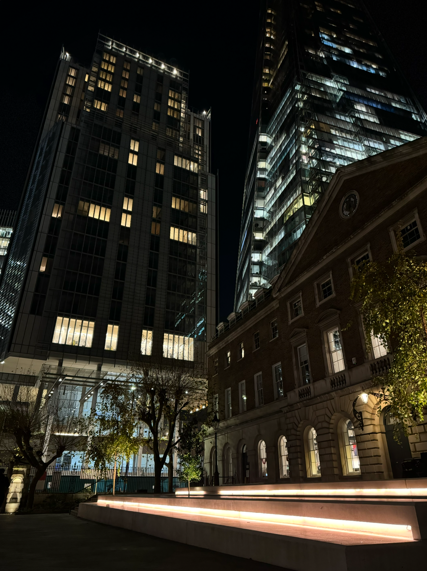 Nighttime city street scene with tall modern glass skyscrapers and an historic brick building, illuminated trees, and stairs with light strips.