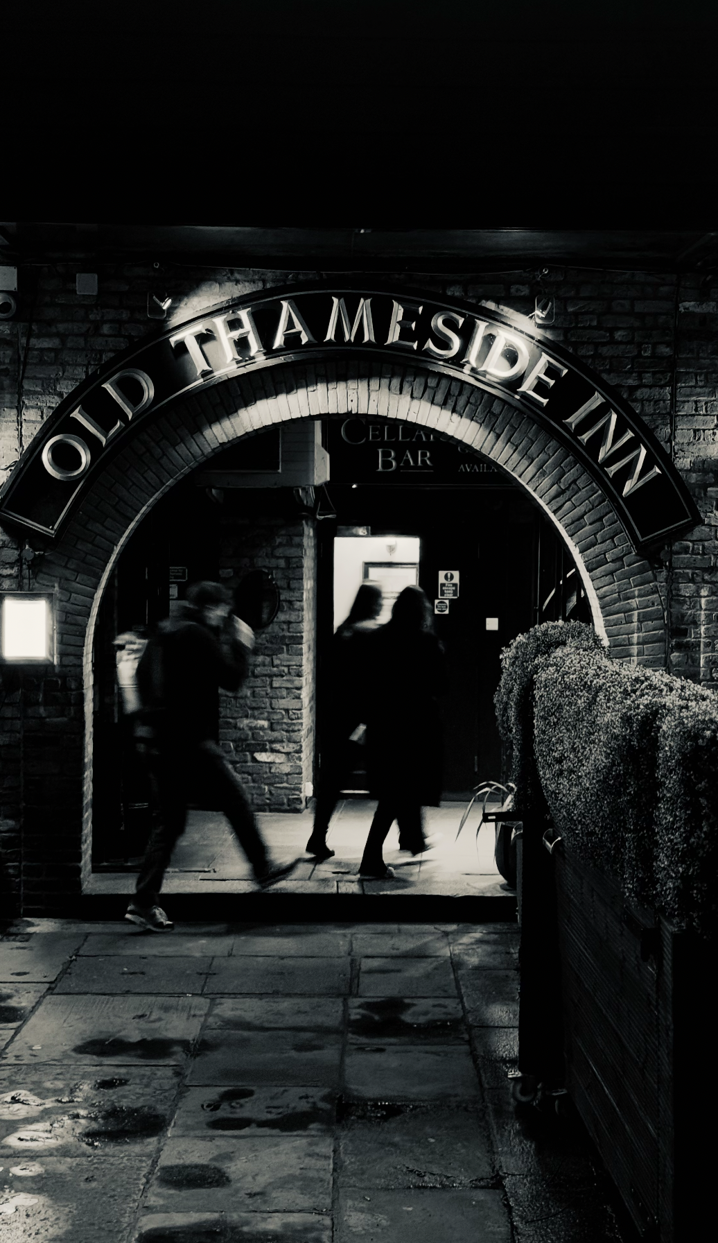 People walking past the entrance of Old Thameside Inn, a brick building with an archway and illuminated sign. Two men and one woman are visible, with one man holding a drink. The scene is dark, suggesting nighttime.