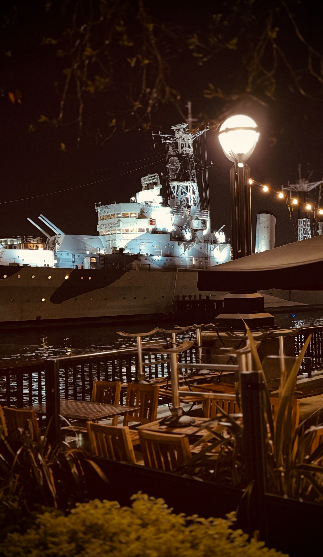 Night scene of a historic battleship docked at a harbor with outdoor seating in the foreground, illuminated by street lamps and string lights.