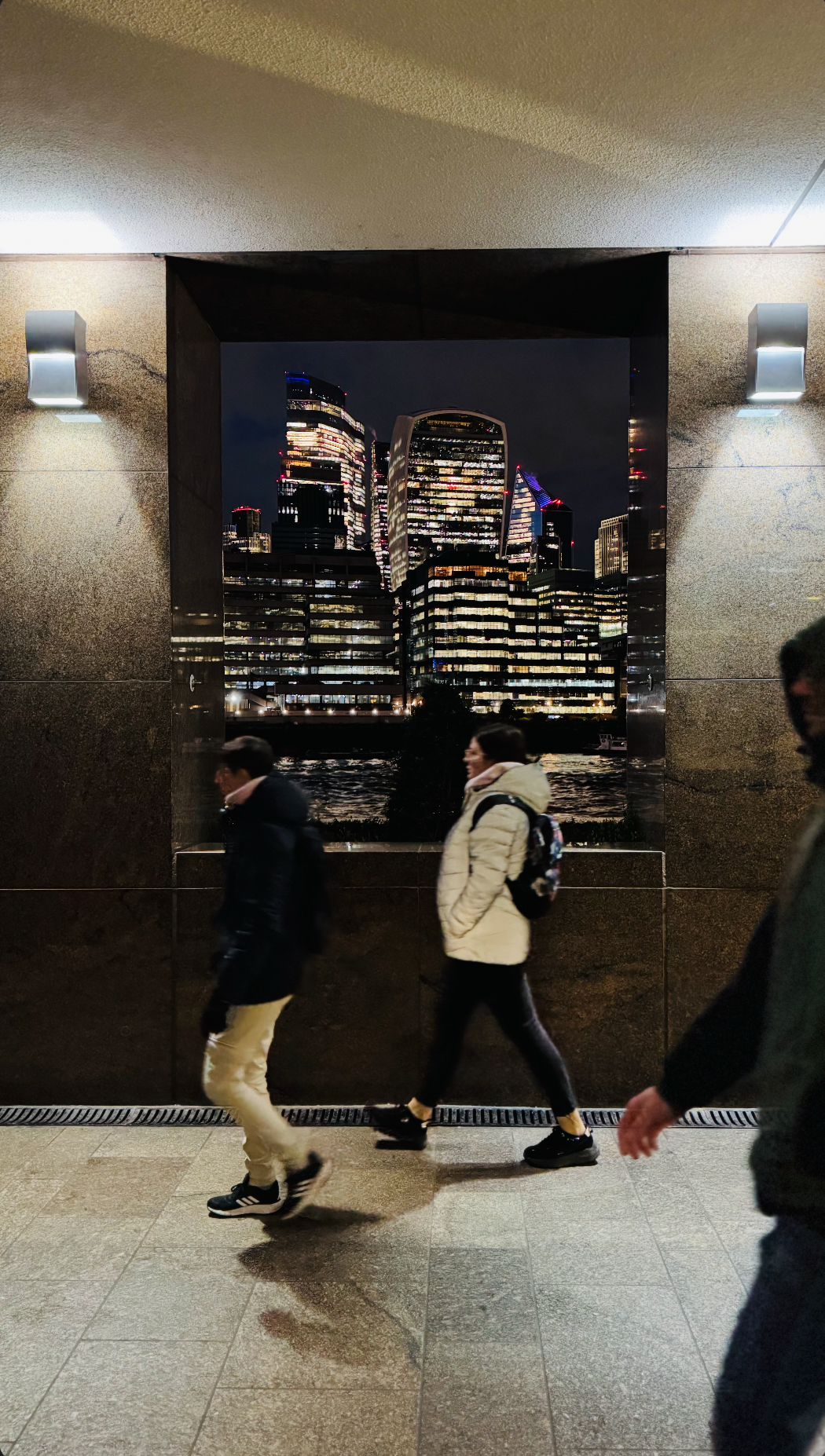 People walking past an urban cityscape photograph of modern illuminated skyscrapers at night, displayed on a wall inside a building.
