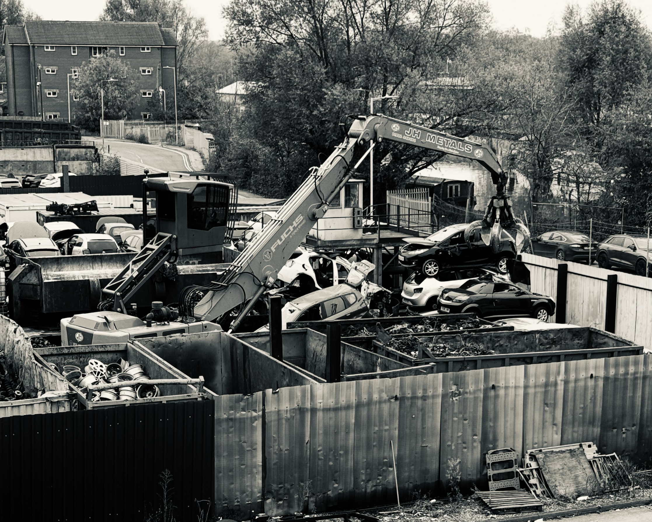 A scrapy yard with a large crane lifting a damaged car, surrounded by stacked and discarded vehicles and car parts.