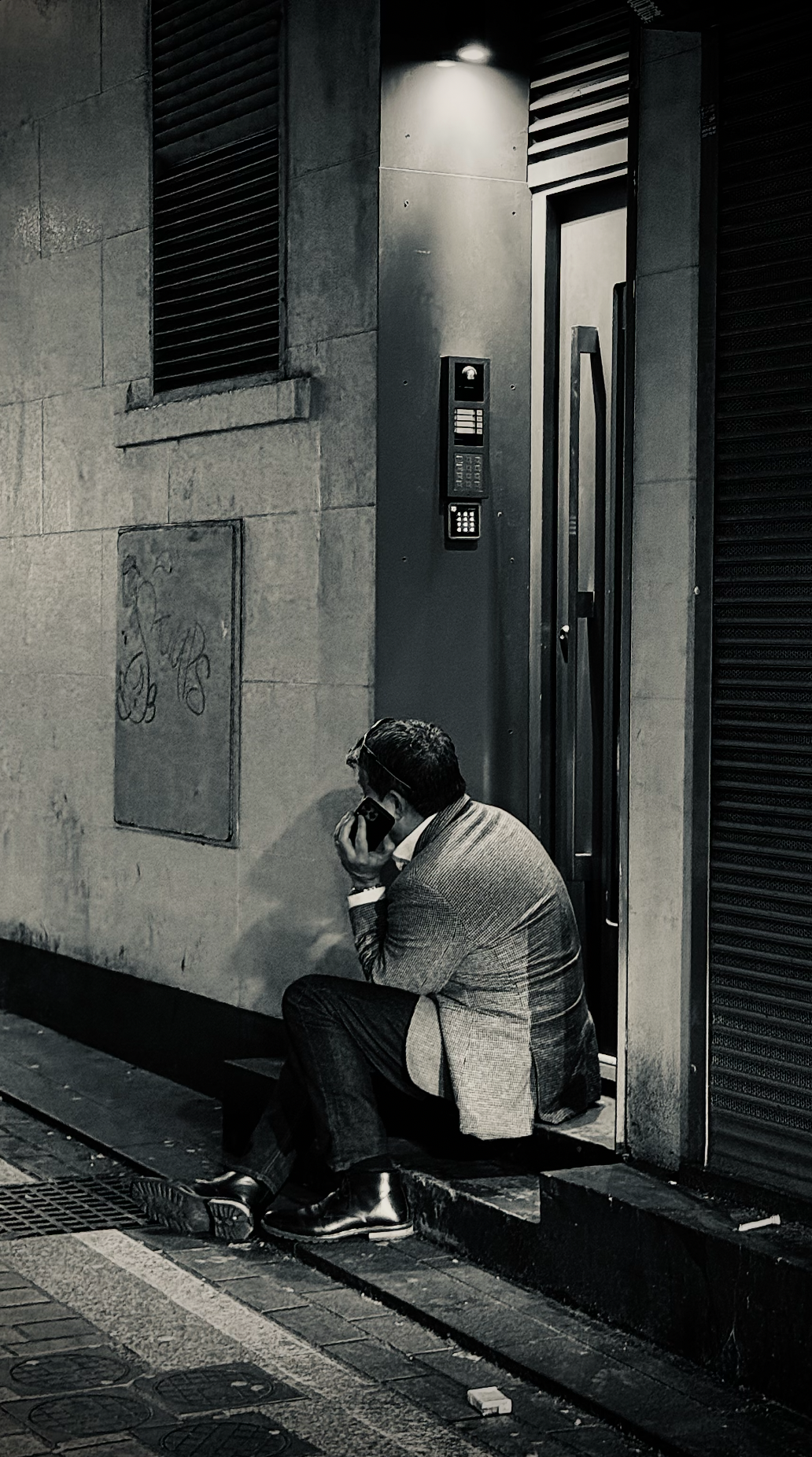 A man wearing a suit and dress shoes sitting on a sidewalk curb outside, talking on a cell phone near a building with intercom and concrete walls.
