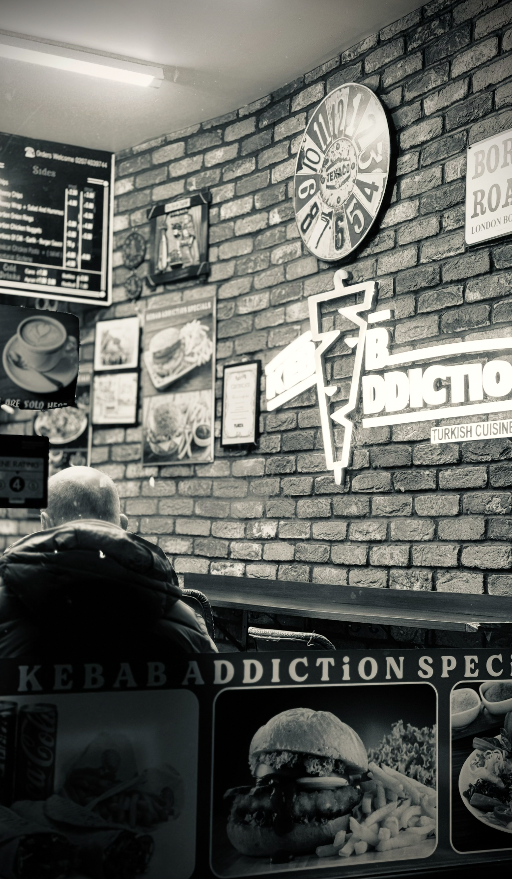 Interior of a restaurant with brick walls, neon sign reading 'Kebabs Addiction Turkish Cuisine,' a clock, various menu boards, and a man sitting with his back facing the camera.