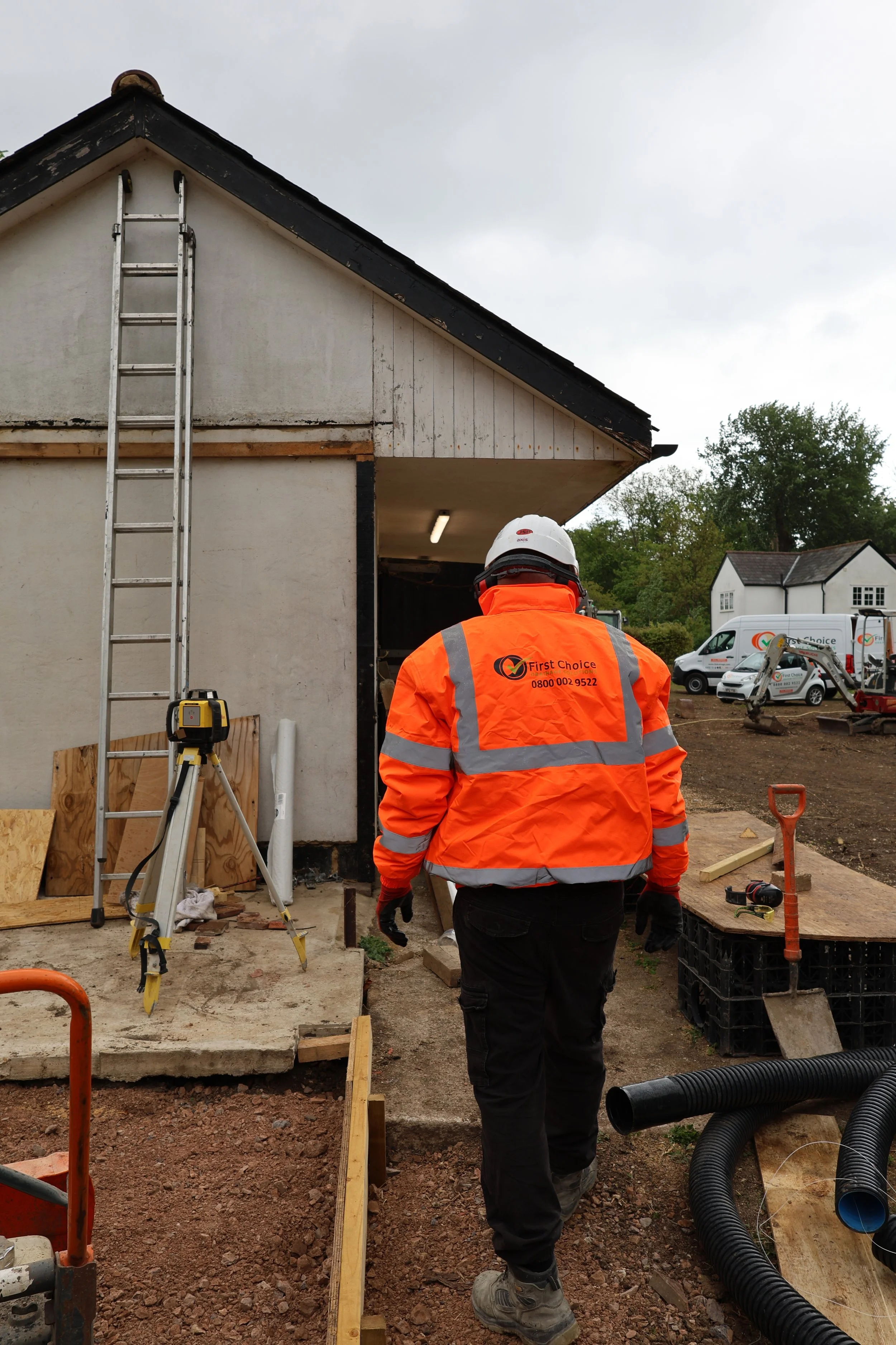 A construction worker in an orange safety jacket walking towards a building under renovation, with construction tools and equipment around.