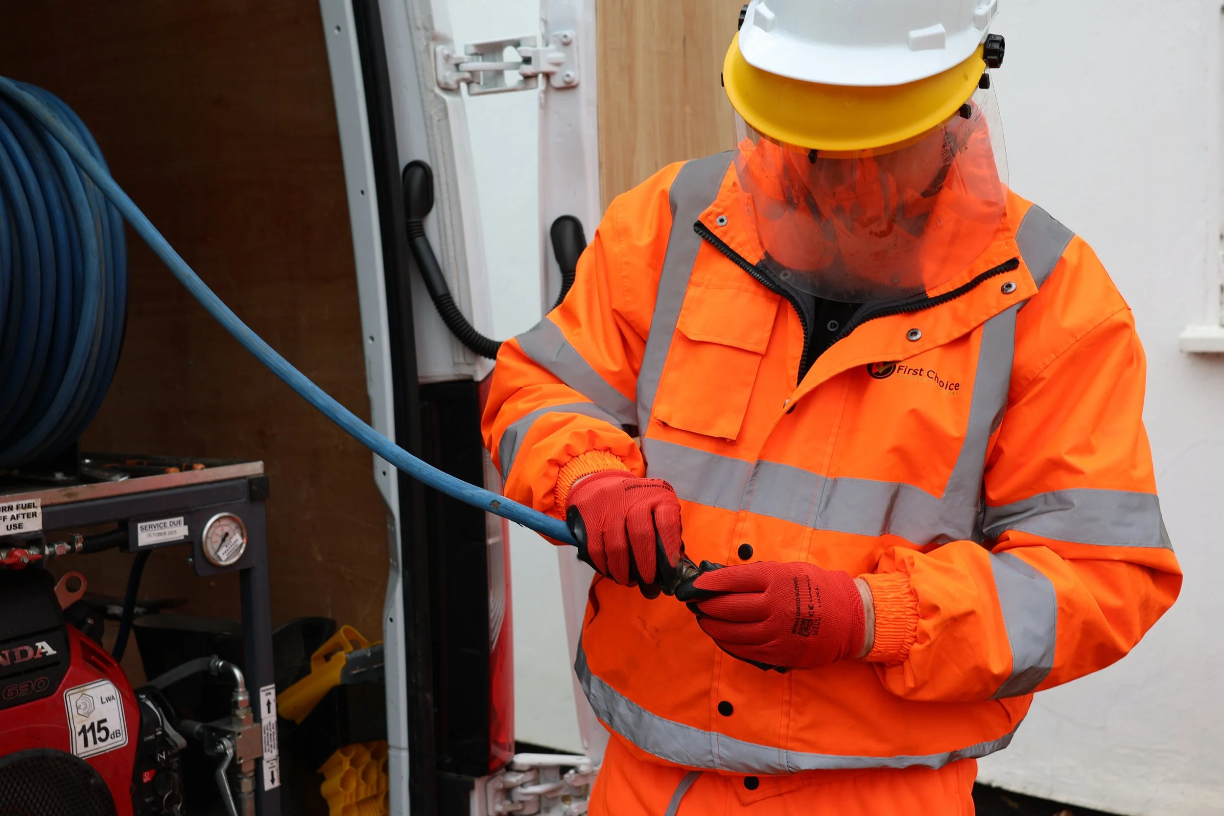 A worker in high-visibility orange safety gear, gloves, helmet, and face shield working with a blue hose near equipment.