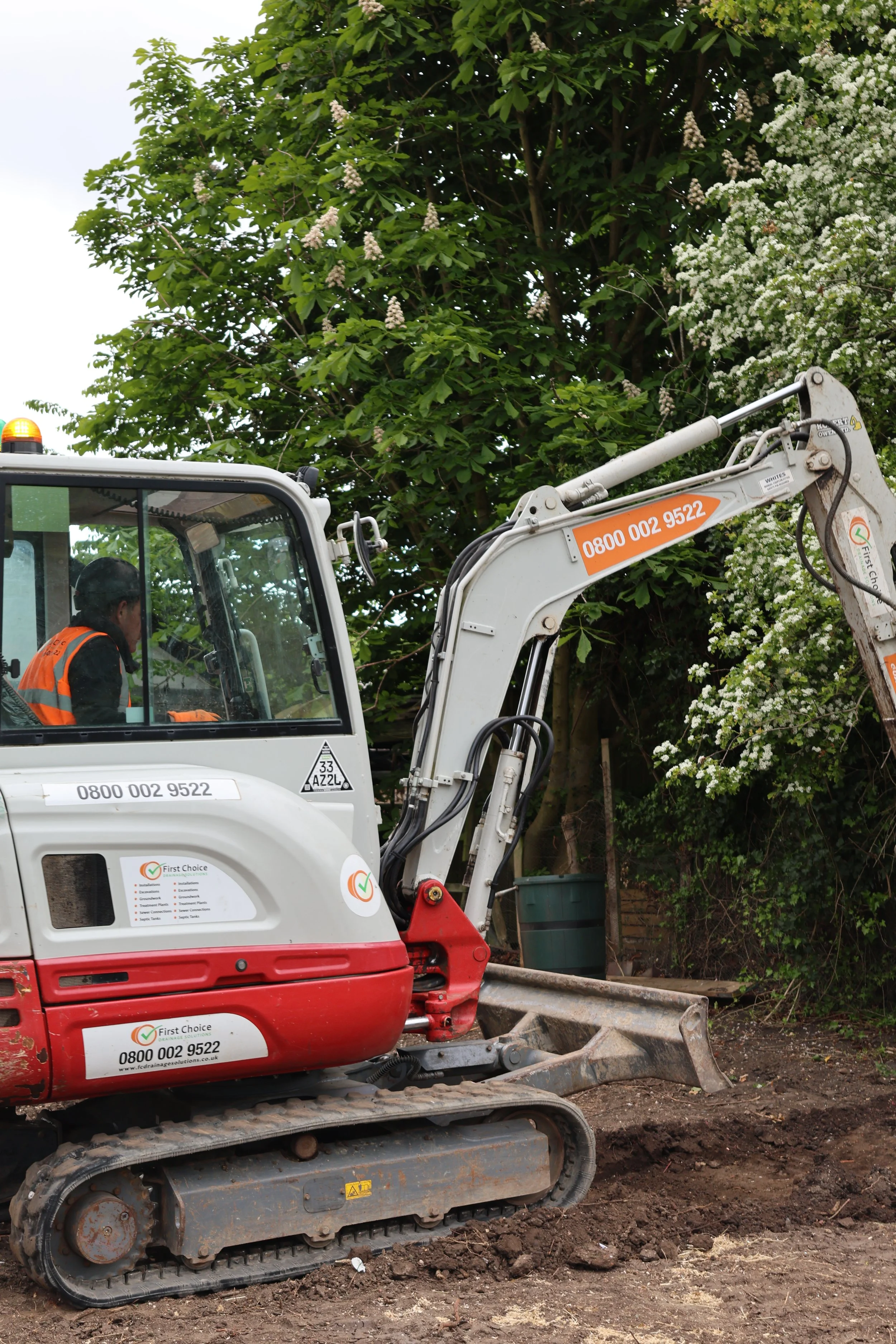 A construction worker operating a small excavator on a dirt ground, with trees and white flowering bushes in the background.