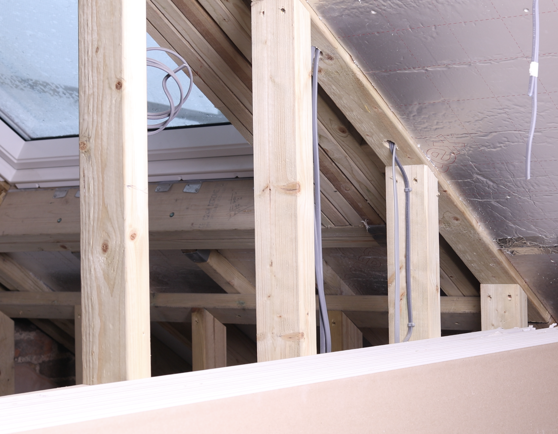 Interior view of an attic under construction with wooden framing, electrical wiring, and insulation installed on the roof.