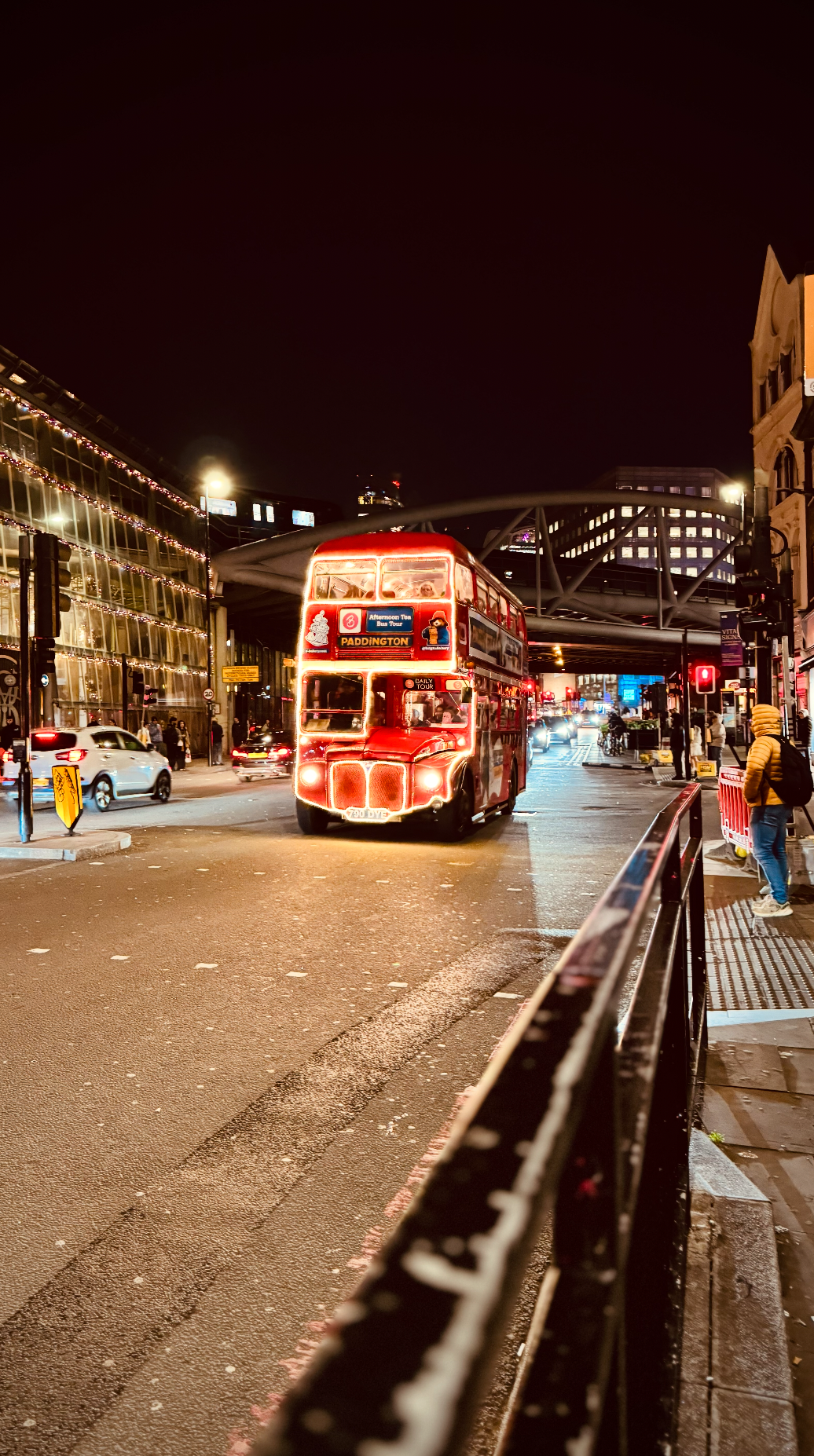 A red double-decker bus at night in a city with modern buildings, illuminated by streetlights, with pedestrians and cars nearby.