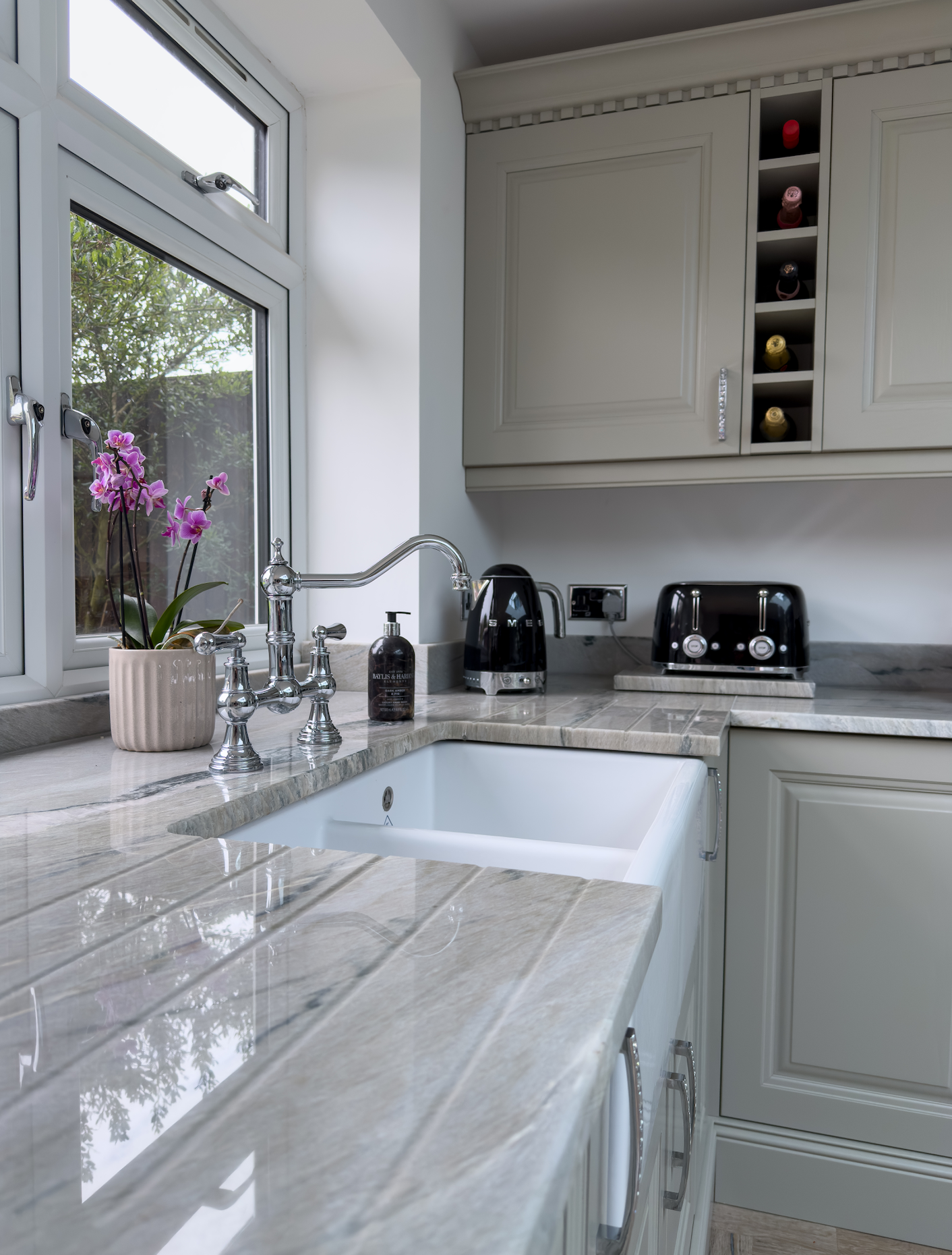 Kitchen with beige marble countertop, white sink, window with view of trees, potted purple orchid, black toaster, black kettle, wine rack in cabinetry.