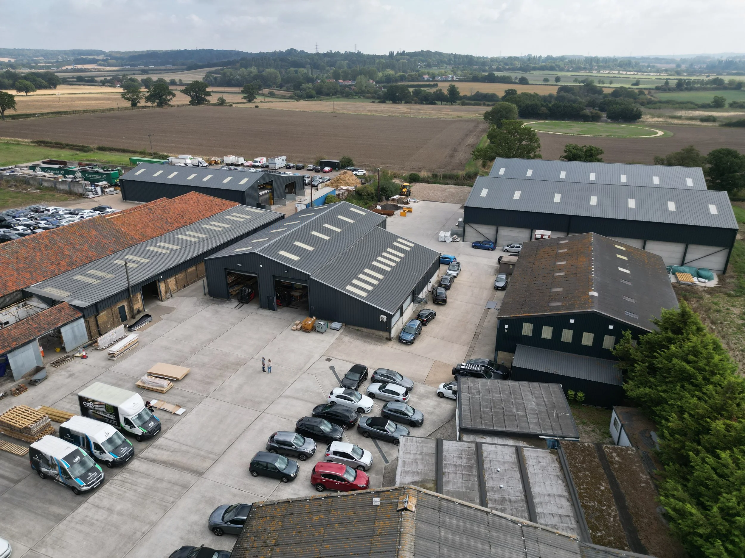 Aerial view of an industrial warehouse complex with multiple black metal buildings, parking lot, and surrounding farmland.