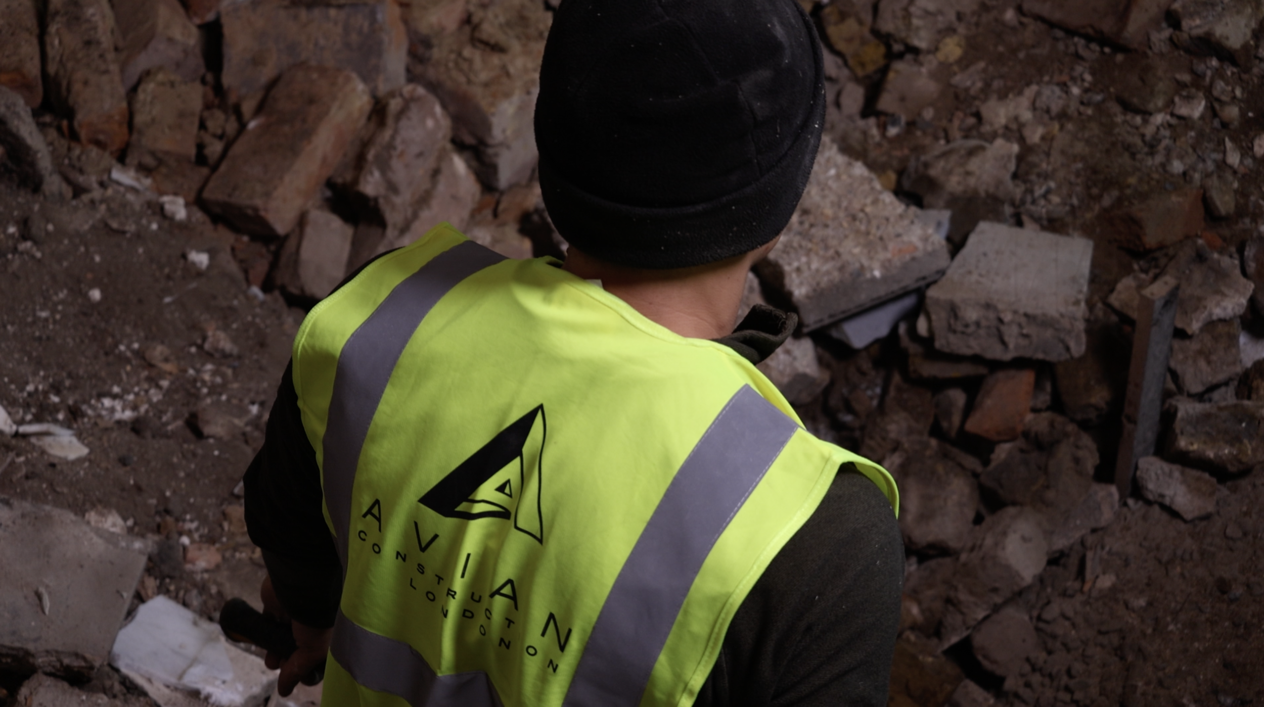 A construction worker wearing a black beanie and a bright yellow safety vest with the logo and name of AVIAN Construction, working amidst a pile of bricks and rubble.