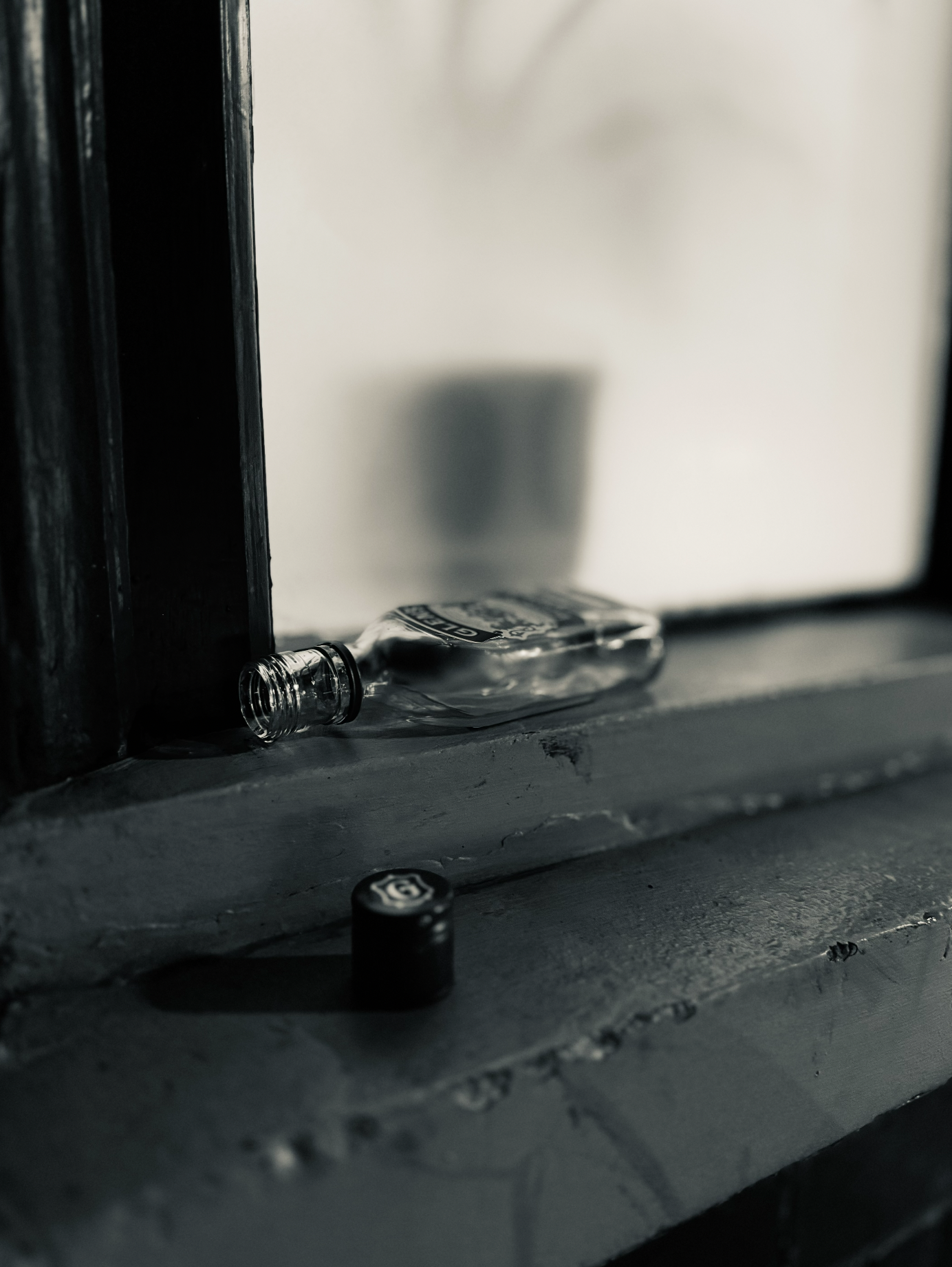 A small glass bottle laying on its side on a weathered windowsill with a black cap placed nearby. The background includes a mirror and part of a dark window frame, with soft lighting creating subtle shadows.